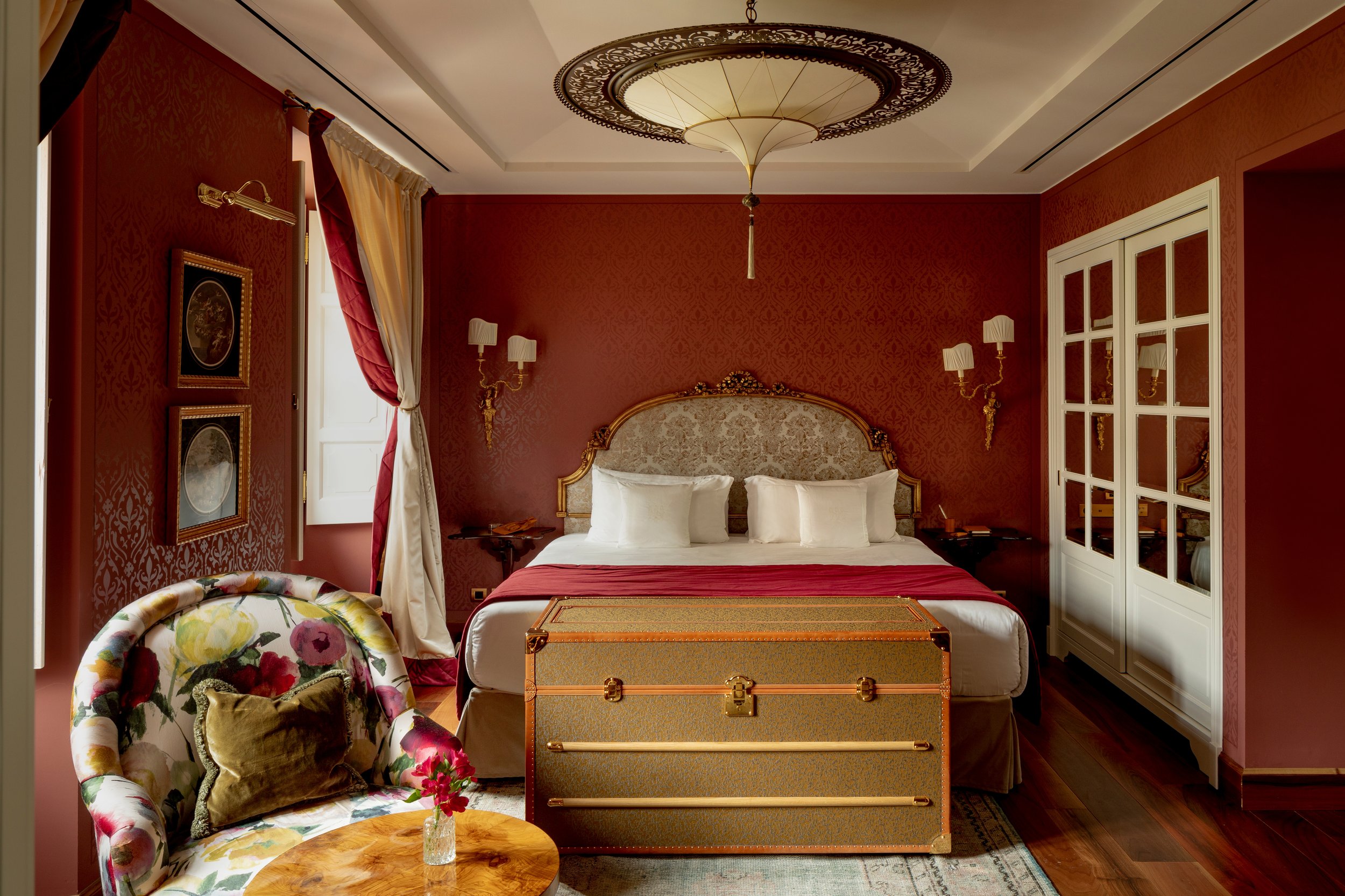 A luxurious bedroom with red damask wallpaper, a king-sized bed with an ornate gold and embroidered headboard, white pillows, and a red bed runner. There are two gold wall sconces with white shades on either side of the bed. To the right is a white wardrobe with glass-paneled doors. To the left, a window with cream and red curtains, and a floral upholstered armchair with a gold pillow. In front of the bed, there is a decorative trunk with a gold finish. A small round wooden table with a vase of flowers and a patterned rug are in the foreground. A large ceiling light fixture with an intricate design hangs from the ceiling.