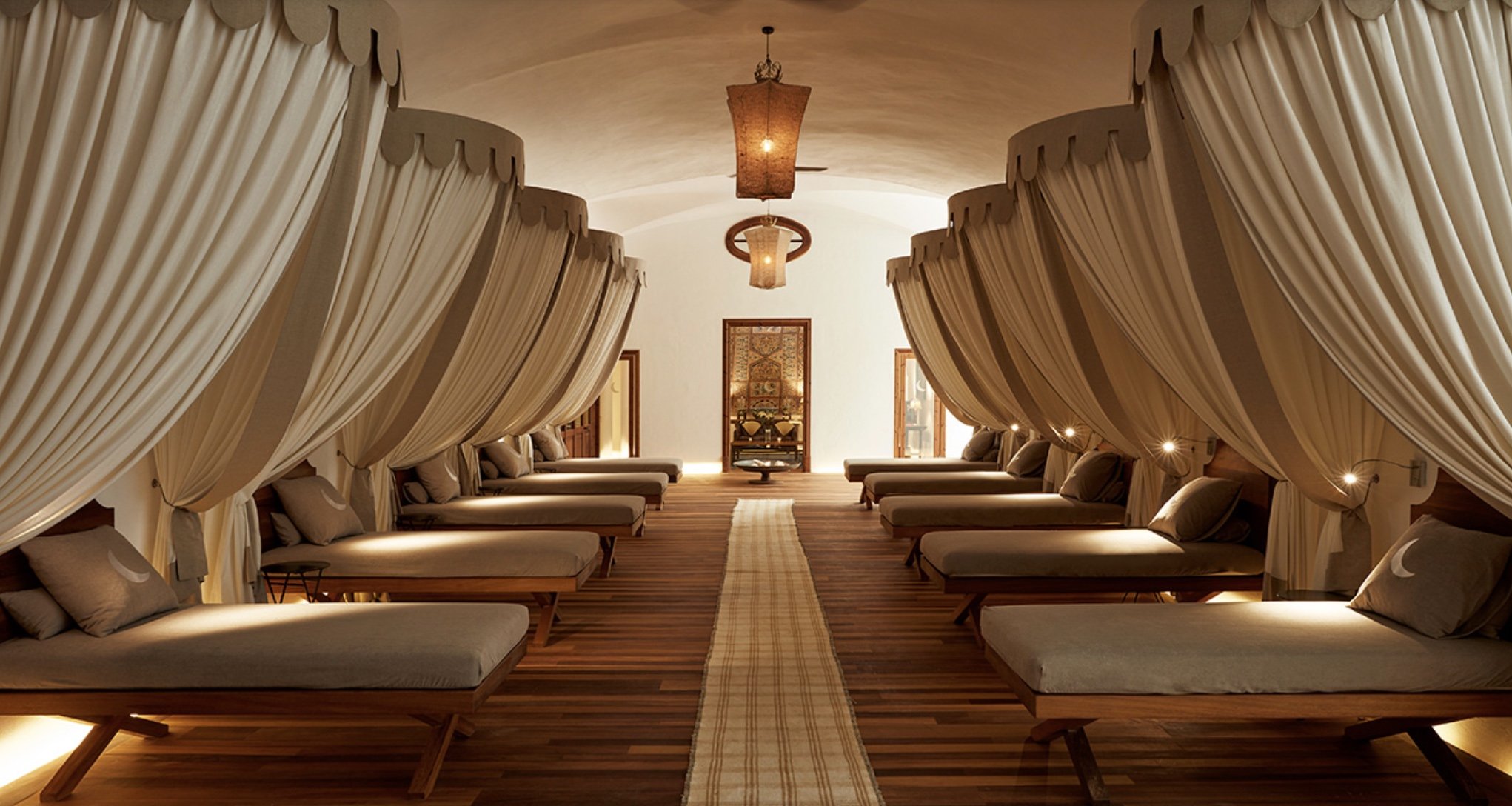 Luxurious spa room with eight bed-like lounge chairs arranged in two parallel rows, each with beige curtains, soft pillows, and dim lighting. A long runner rug runs down the center, with decorative lamps hanging from the ceiling.