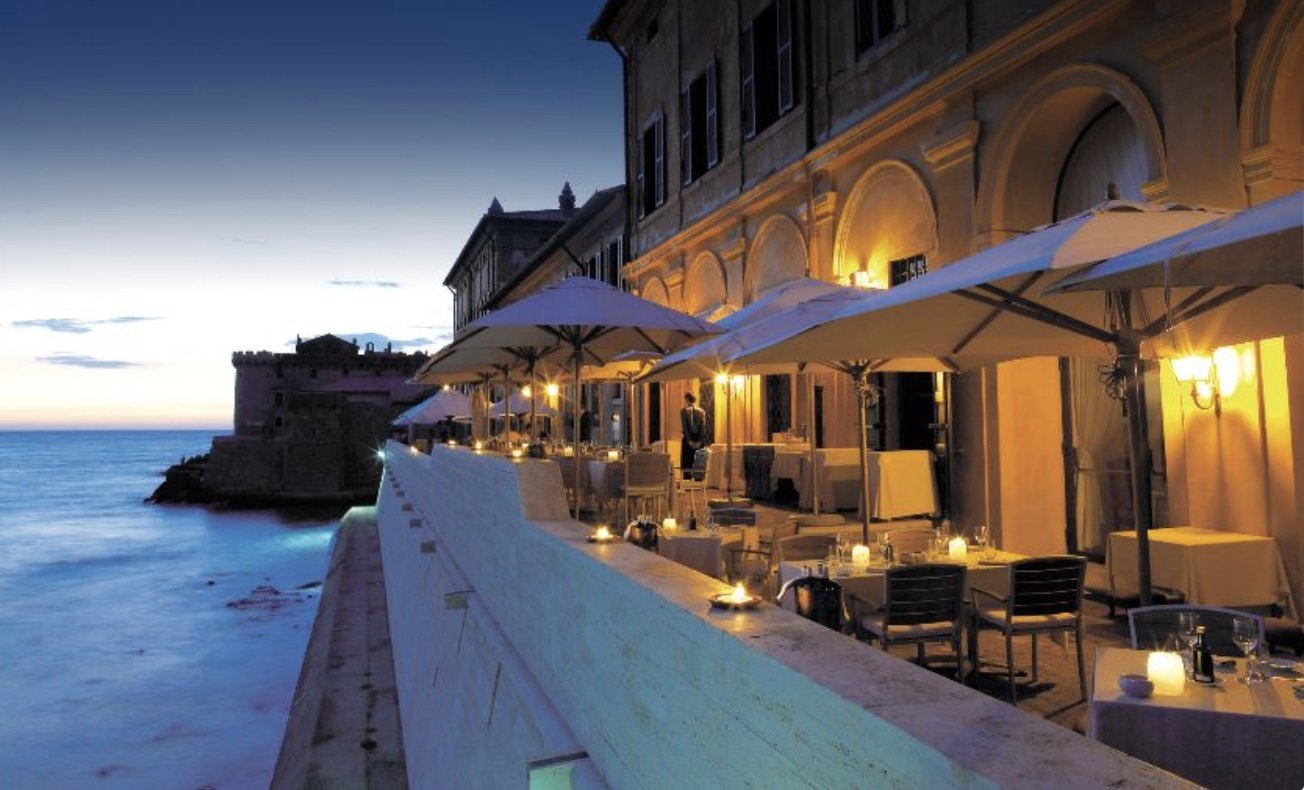 Outdoor dining area with tables, chairs, and umbrellas illuminated by warm lighting, overlooking the ocean at sunset or dusk, with historic buildings in the background.