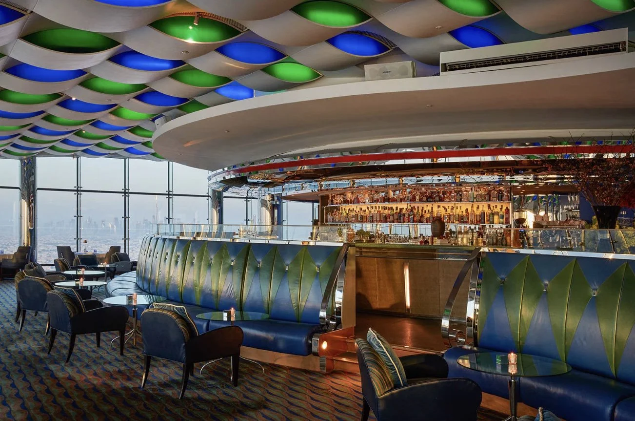 Interior of a stylish bar with a curved seating area, high-backed blue and green upholstery, and a well-stocked bar with bottles illuminated in the background. The ceiling features a pattern of recessed, colored lights, and large windows offer a city