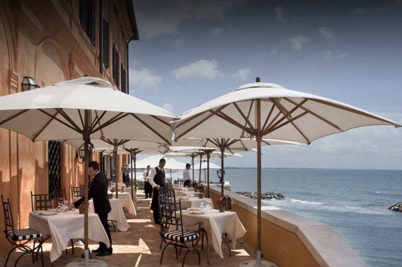 An outdoor seaside restaurant with tables covered in white tablecloths, black and white striped chairs, and large white umbrellas providing shade, overlooking the ocean with a partly cloudy sky.