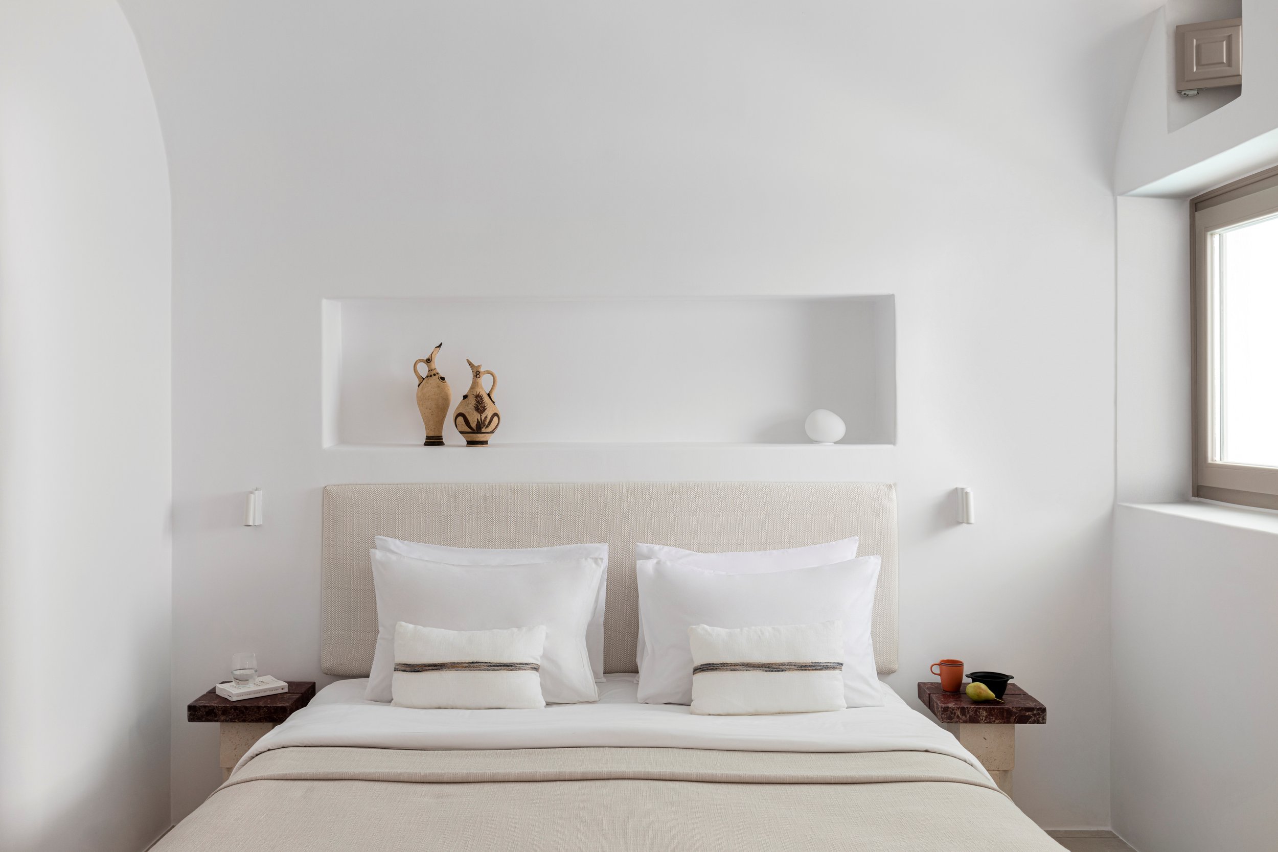 A minimalistic bedroom with a white bed, beige headboard, and two pillows. On the left nightstand is a glass of water and some items, and on the right are a mug, a bowl, and a pear. The wall behind the bed features a recessed shelf with two decorativ