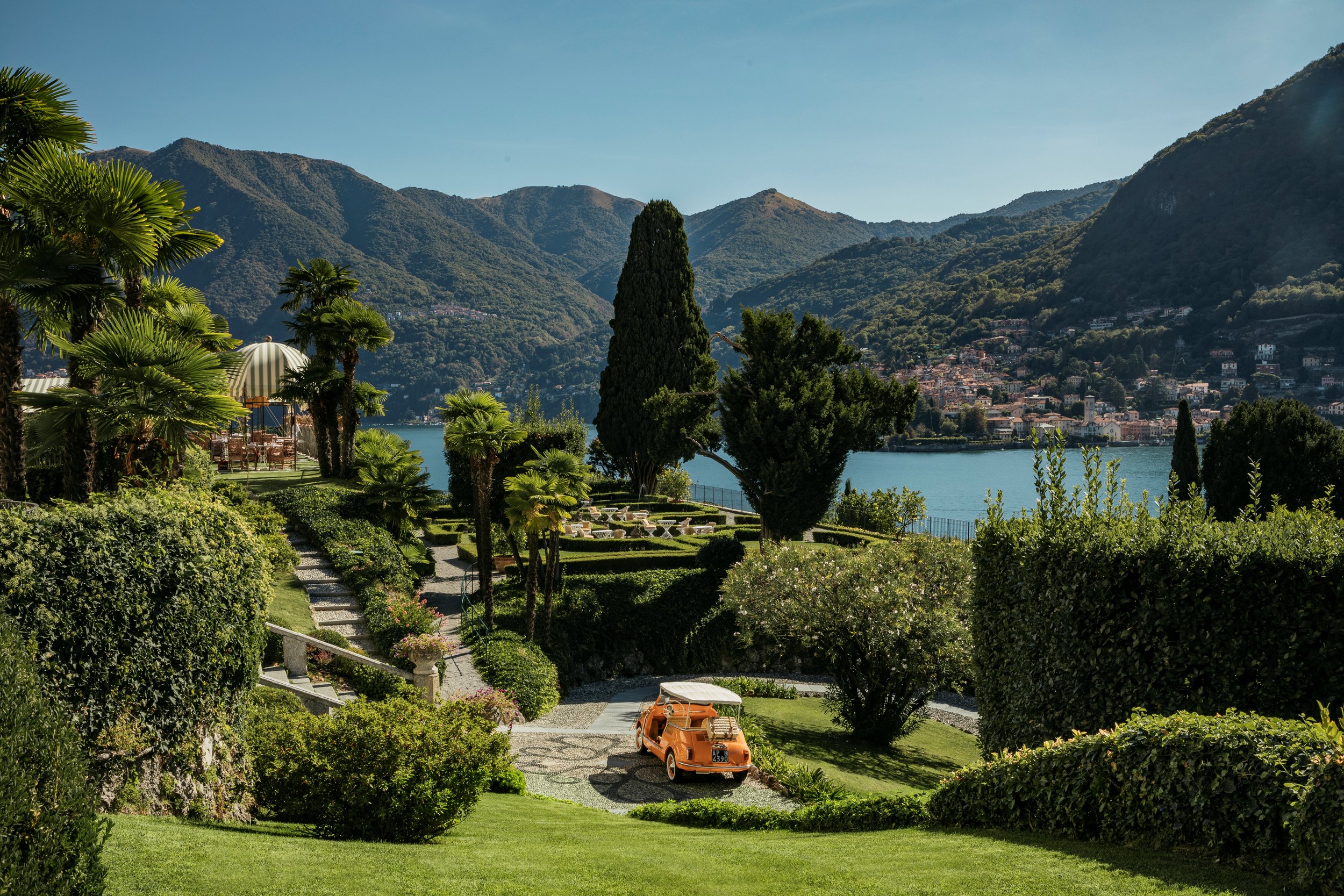 A lush, landscaped garden with a vintage orange car parked on a gravel driveway, overlooking a lake with a small town and mountains in the background.