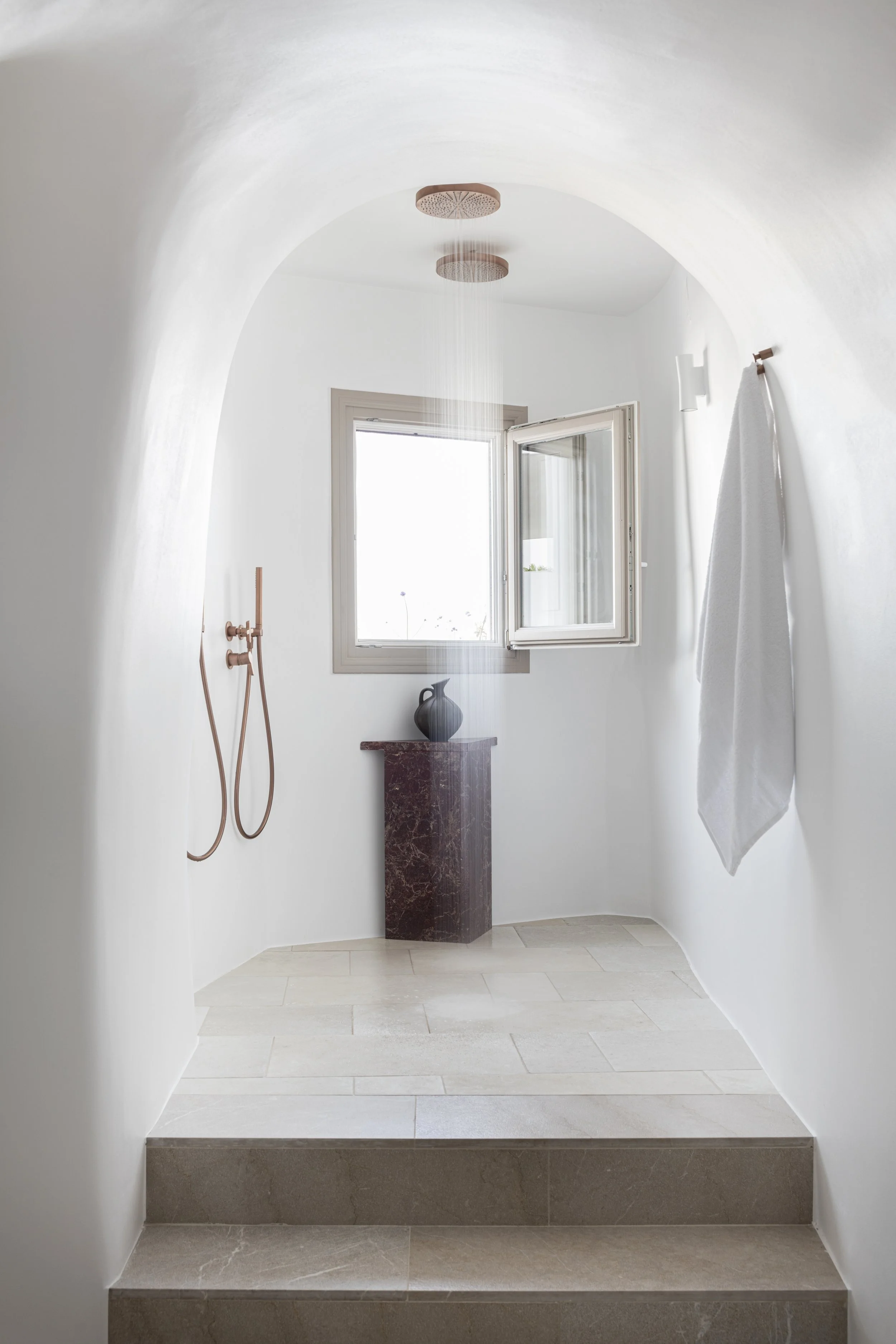 Minimalist walk-in shower with two overhead rain shower heads, an open window, a small dark marble pedestal, and a white towel hanging on a wall