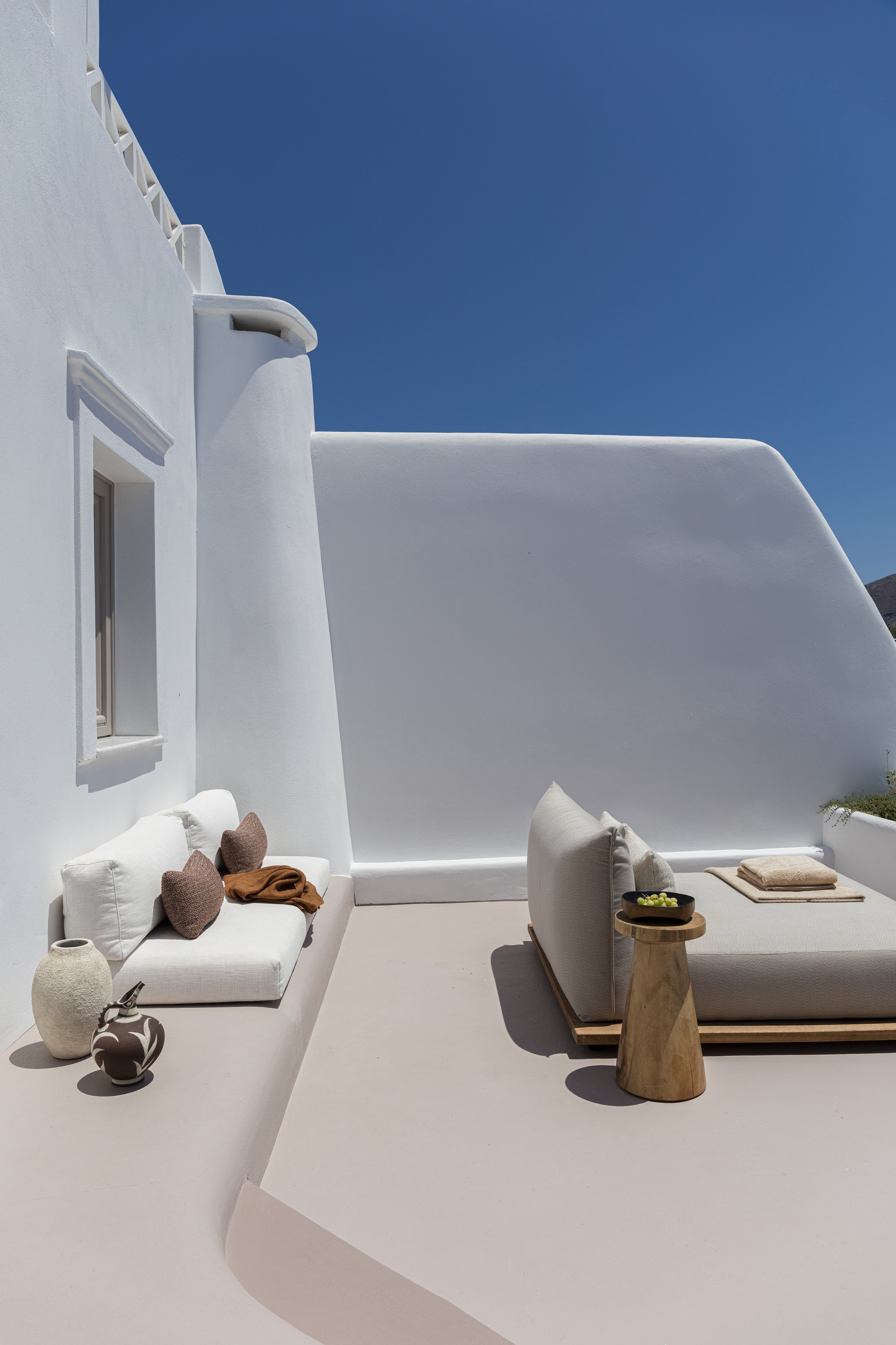 A minimalist outdoor patio with white walls and furniture, featuring a cushioned lounge chair with pillows, decorative vases, and a small side table with grapes, against a bright blue sky.