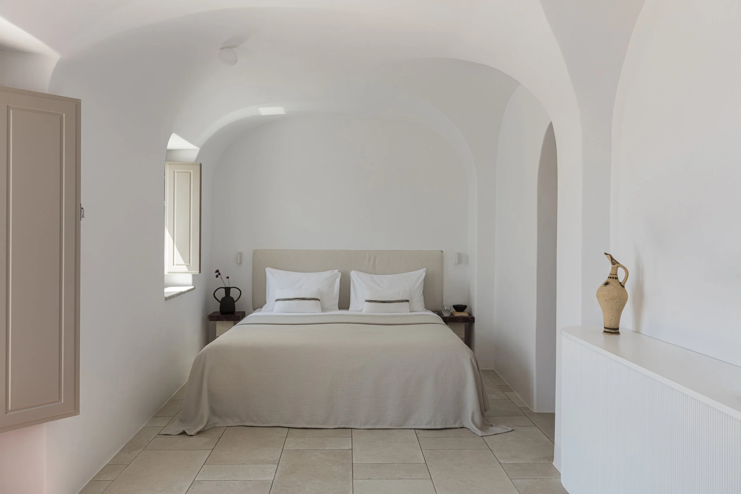 A minimalist bedroom with white walls, a bed with white linens, pillowed headboard, two small bedside tables with a black vase and a cup, a window with open shutters, a beige cabinet, and decorative pottery on a white console.
