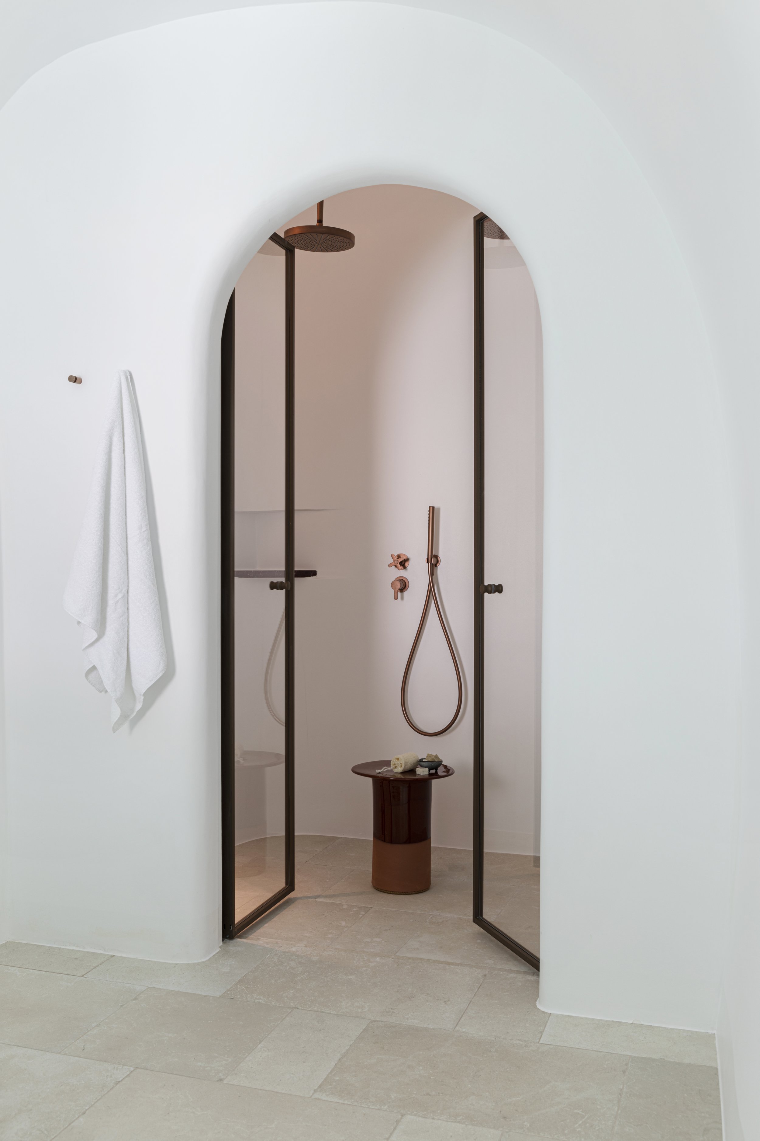 Minimalist shower with a white wall, glass door, rain showerhead, and a brown stool with toiletries inside.