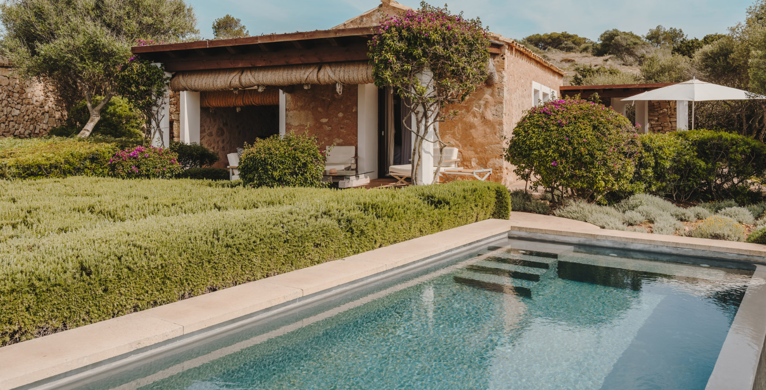 A backyard of a house with a swimming pool, green bushes, purple flowers, trees, and a patio with outdoor furniture and a sunshade.