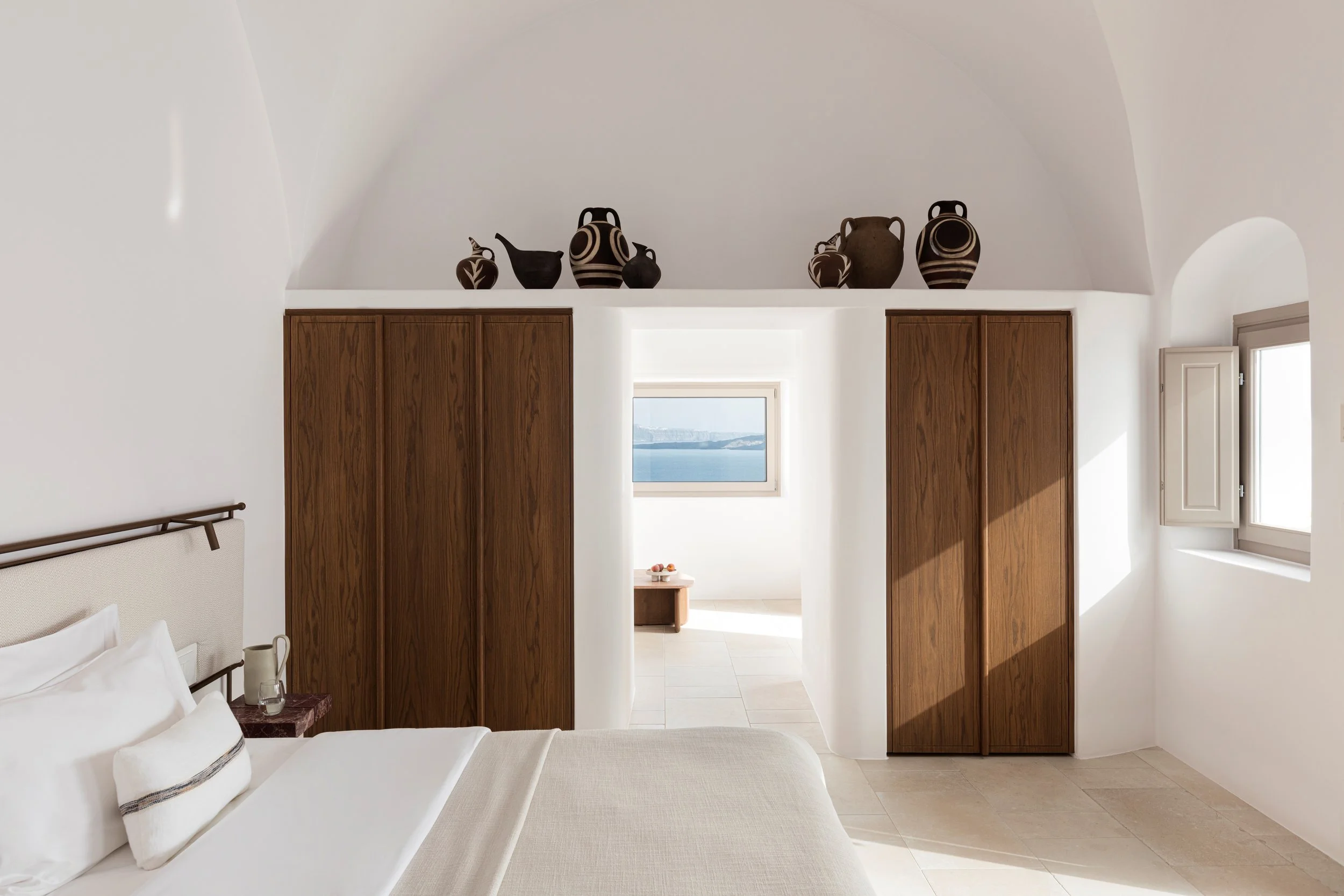 A bedroom with a white bed, beige pillows, wooden closet doors, a small wooden nightstand, a window with a view of the ocean, and decorative black and brown vases on a high shelf.