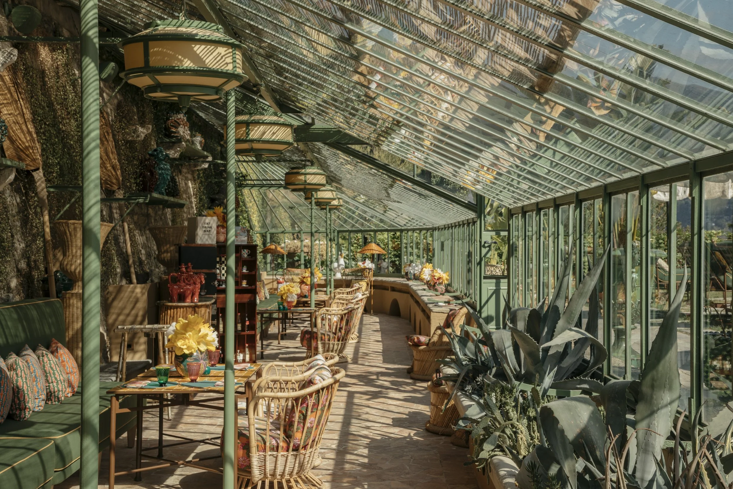 Sunlit greenhouse with green metal framing, containing various wicker chairs, plants, flowers in vases, and decorative items.