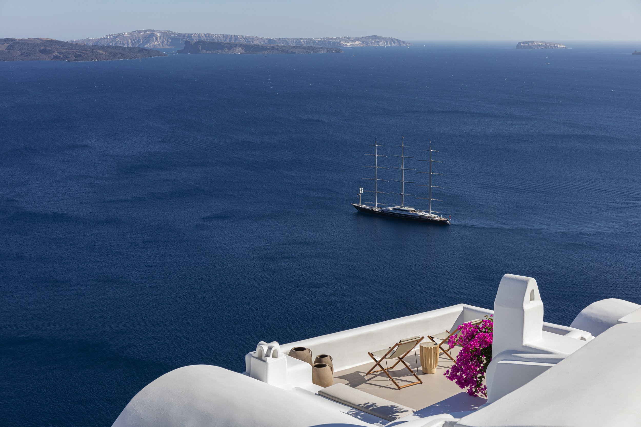 View of the Aegean Sea from a white terrace with pink flowers and beach chairs, with a sailing ship in the water and islands in the distance.