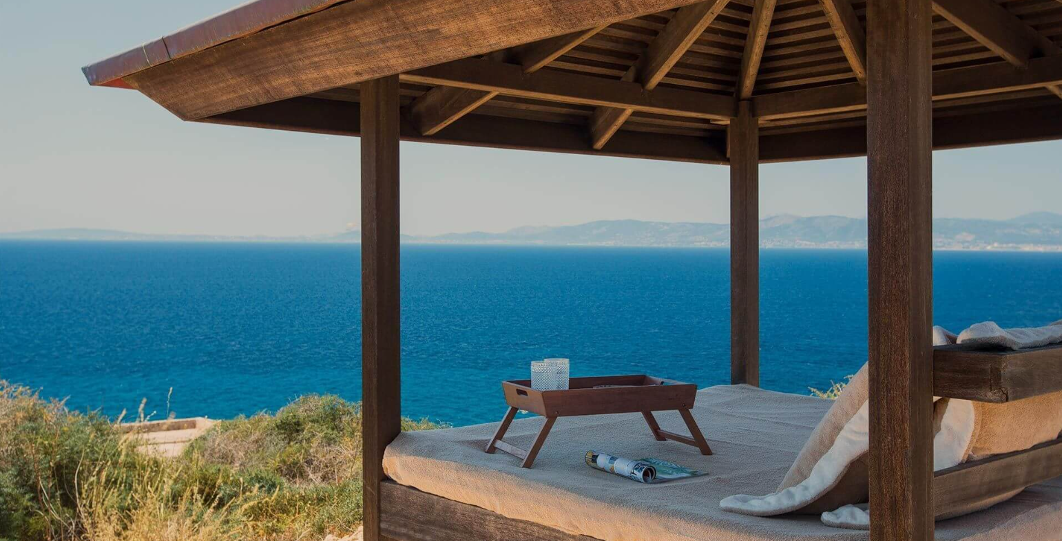 Beachside cabana with a canopy, overlooking the ocean with hills in the distance, with a tray, rolled magazine, and cushions inside.