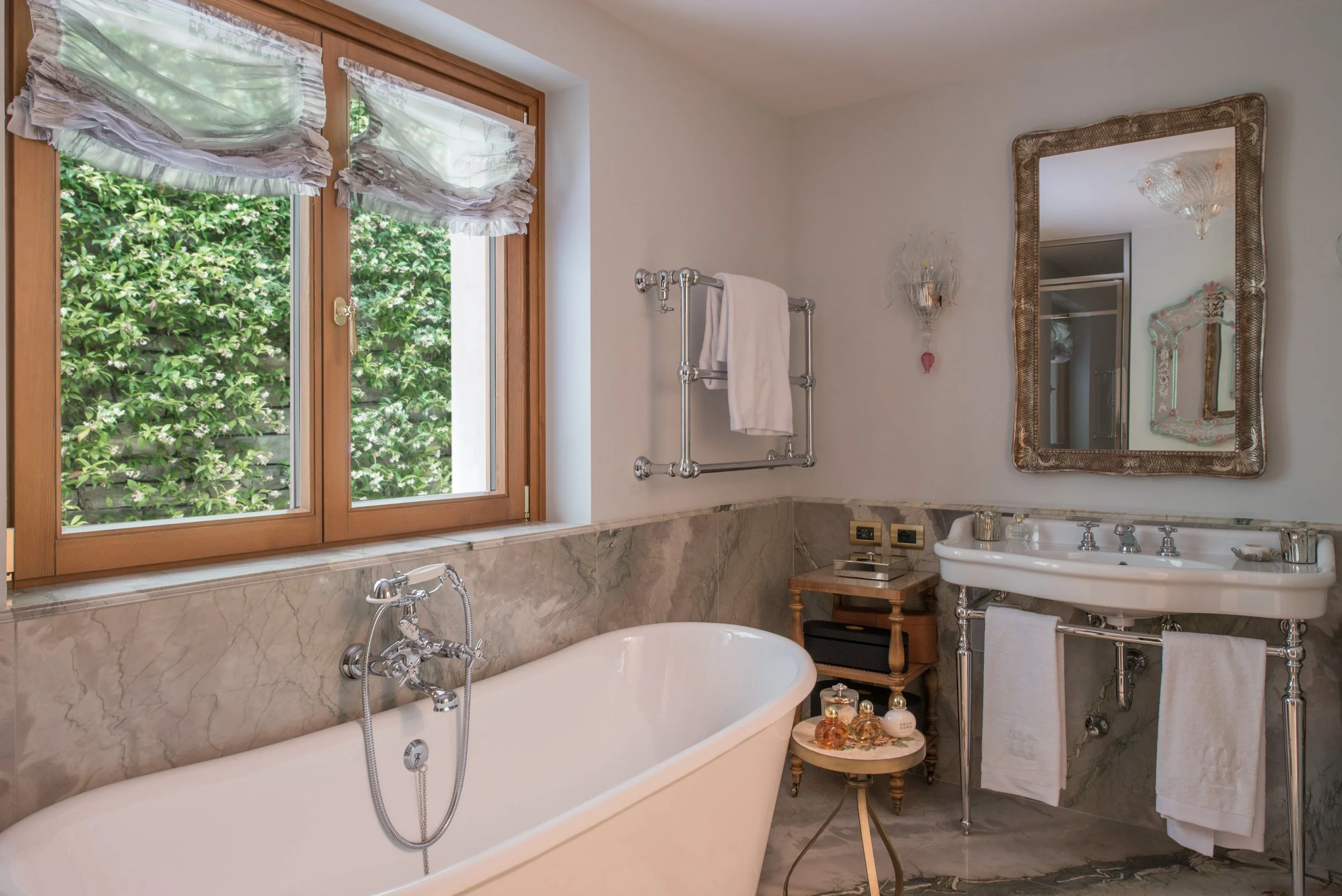Bathroom with a large bathtub below a window with floral curtains, a marble-surfaced sink with towel racks, and a mirror with ornate frame, a part of chandelier, and a small side table with perfume bottles.