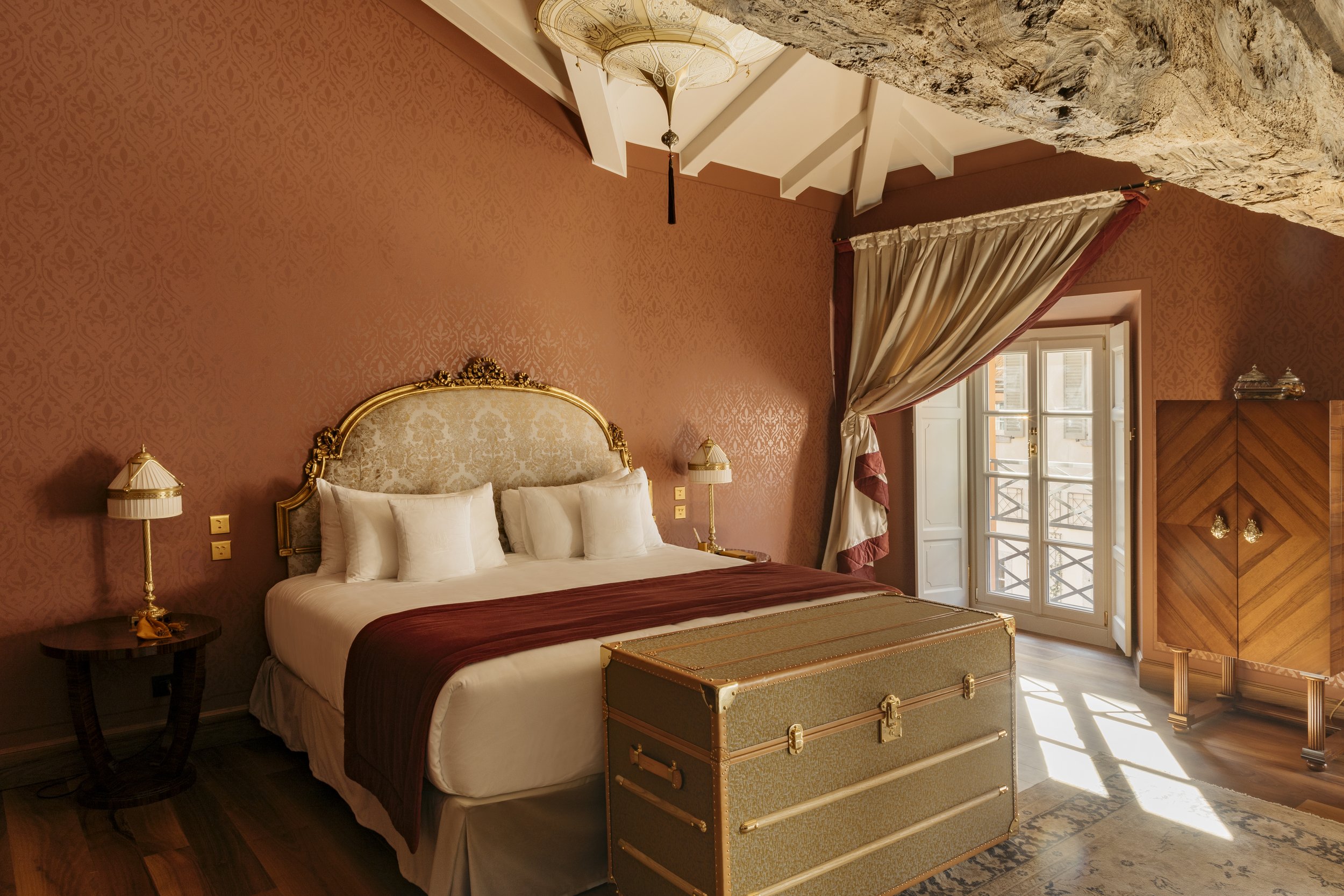 A luxurious bedroom with a large bed featuring white pillows, a decorative gold headboard, and a maroon bedspread. The room has pinkish patterned walls, a wooden side table with a lamp on each side, a window with cream-colored curtains, a textured ceiling, and a large chest at the foot of the bed. There is also a wooden wardrobe and a balcony door allowing natural light to enter.