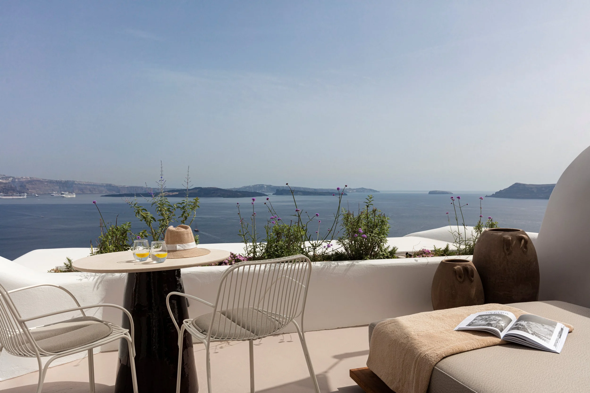 Outdoor patio with white seating area, a small round table with two glasses of yellow drink and a straw hat, overlooking a large body of water with islands in the distance, and potted plants along the white wall.