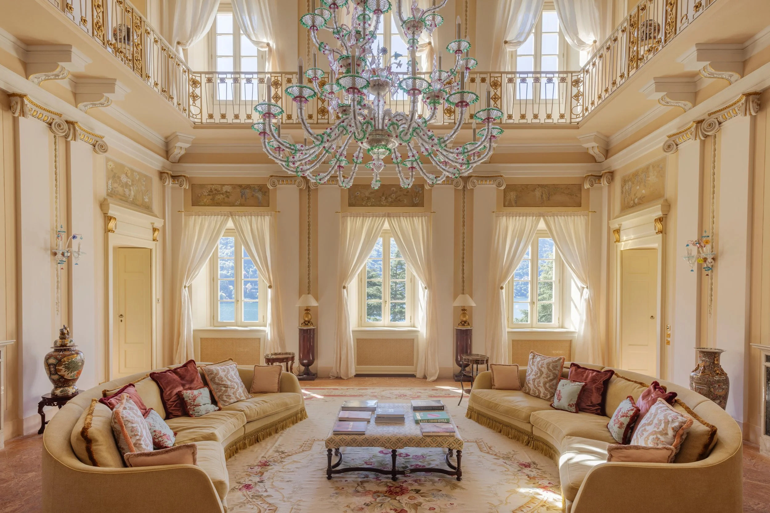 Elegant living room with high ceilings, large windows with cream curtains, a grand chandelier, cream-colored sofas with decorative pillows, a central coffee table with books, and ornate vases and lamps.