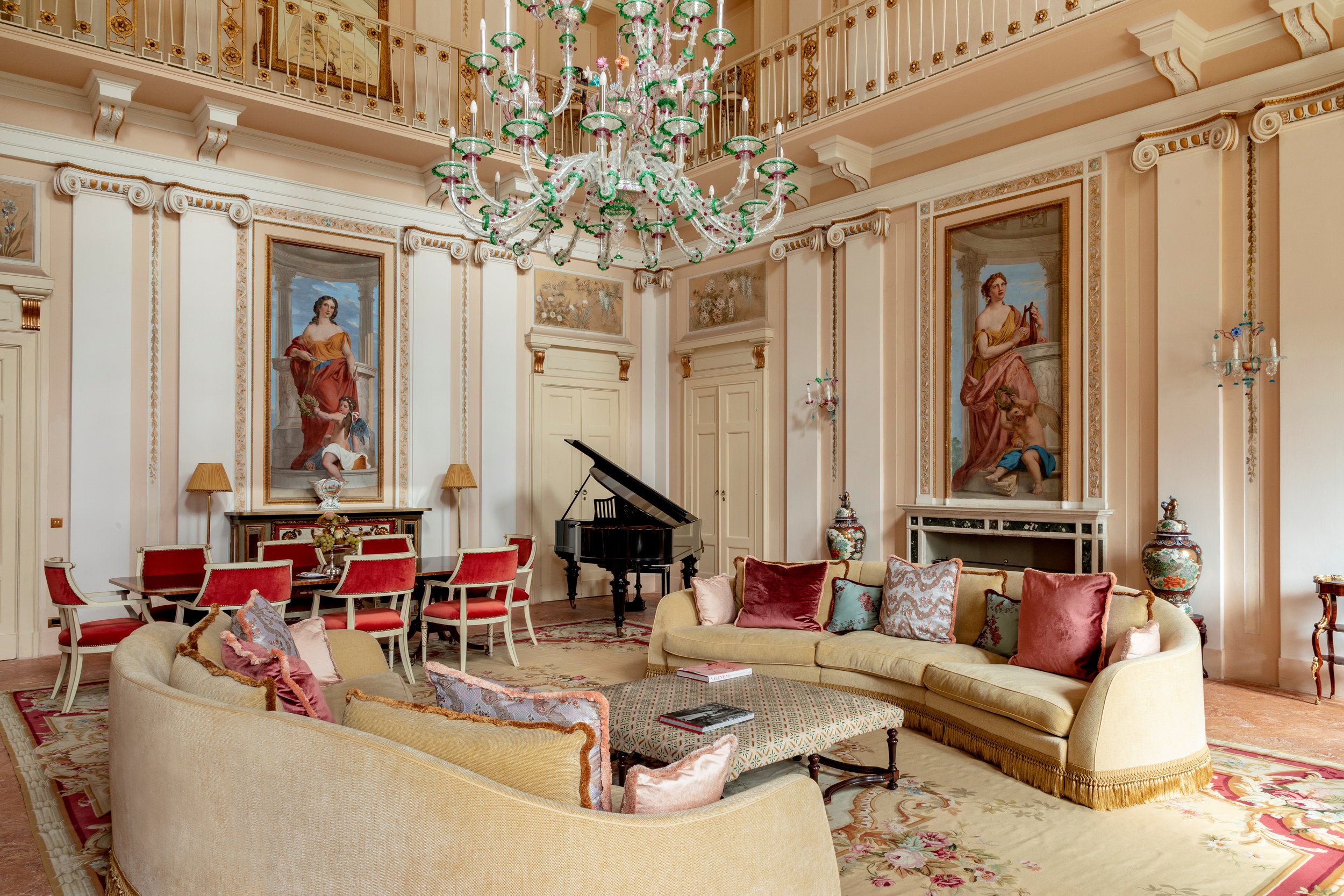 Elegant living room with a grand chandelier, classical paintings, a black grand piano, and luxurious furniture including a cream sofa with colorful pillows and a red dining table with matching chairs.