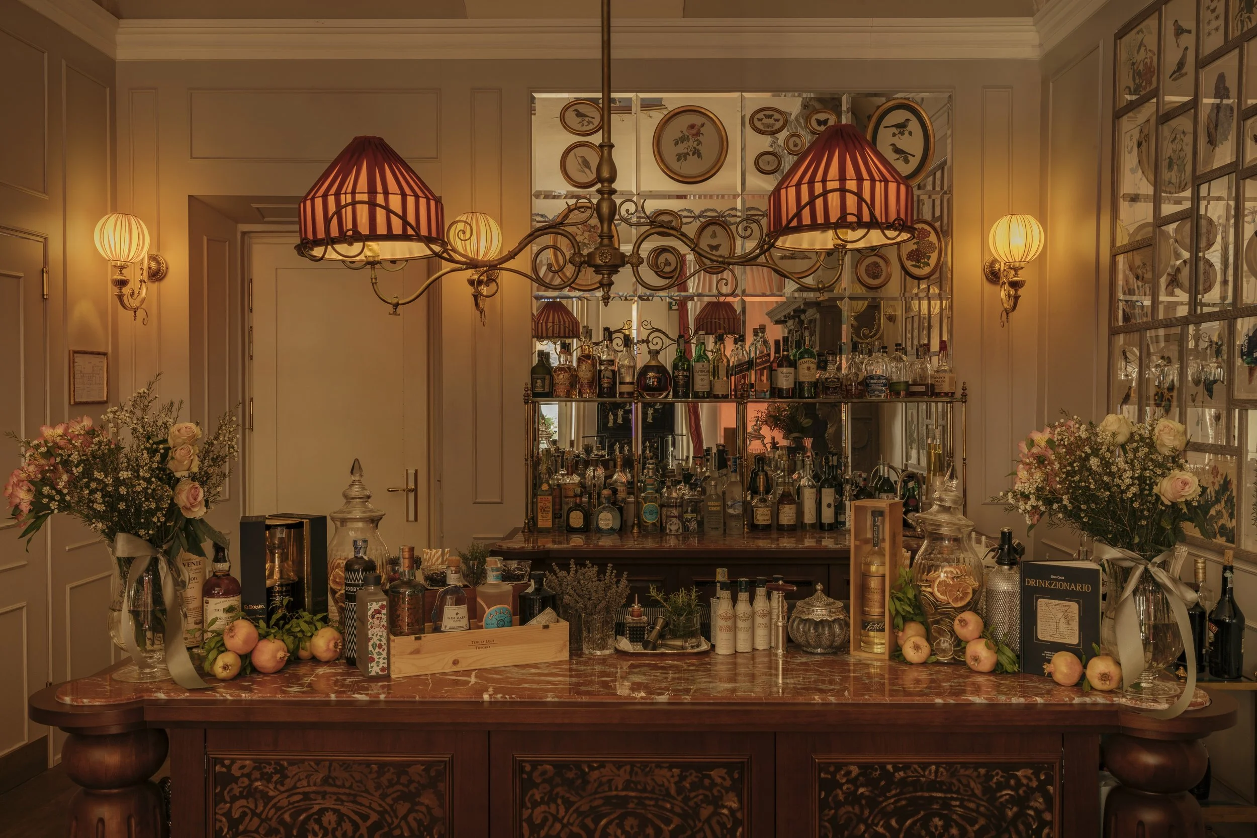 An elegant home bar with a marble countertop, floral arrangements, and a mirror behind displaying various bottles of alcohol. Warm lighting from a chandelier with red lampshades and wall sconces create a cozy atmosphere.