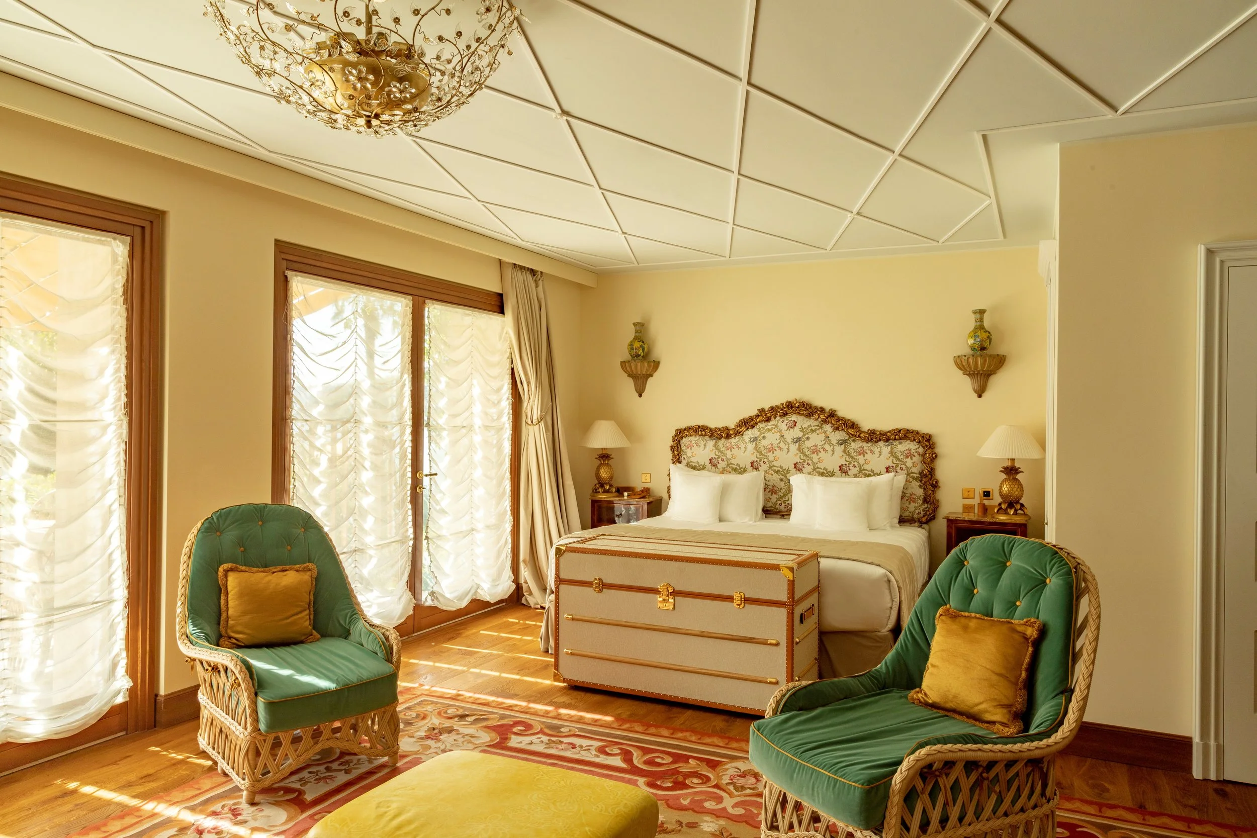 A luxurious bedroom with a bed featuring an ornate floral headboard, two side tables with lamps, armchairs with green upholstery and gold pillows, a chandelier, large windows with curtains, and a trunk at the foot of the bed.