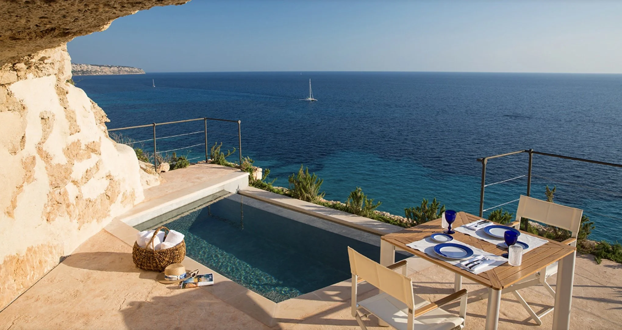 An outdoor patio with a small pool overlooking the ocean. There is a table set with plates, glasses, and utensils, and two chairs. A basket with rolled towels and a sun hat rests near the pool. The scenery includes clear blue water and boats on the h