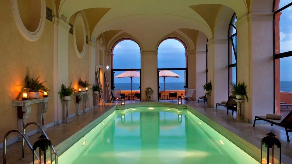 Indoor swimming pool area with large arched windows revealing a view of the ocean and beach umbrellas on the patio outside, decorated with plants, candles, and lounge chairs.