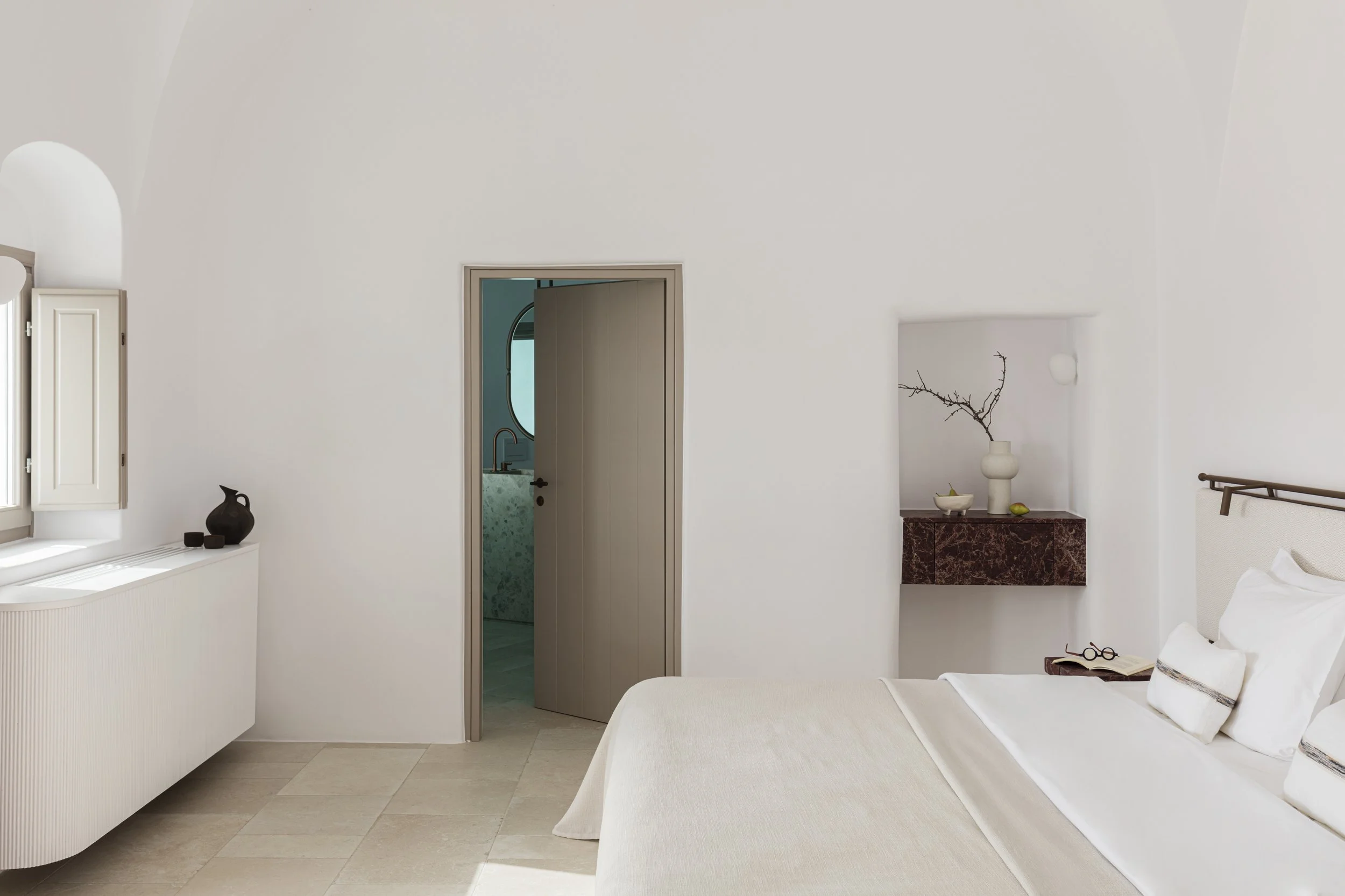 A minimalist bedroom with a white bed, decorative pillows, a small bedside table with glasses and a book, a niche with a vase and branches, a door leading to a bathroom, and a window with shutters.