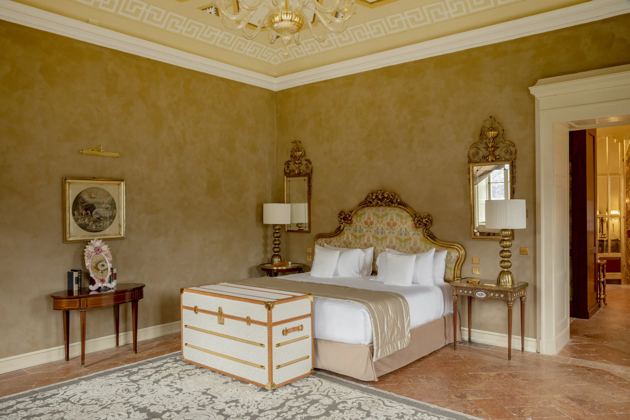 Luxurious hotel bedroom with a large ornate bed, matching nightstands, and tall table lamps. The room has beige walls, decorative mirrors, framed artwork, a chandelier, and a patterned area rug.