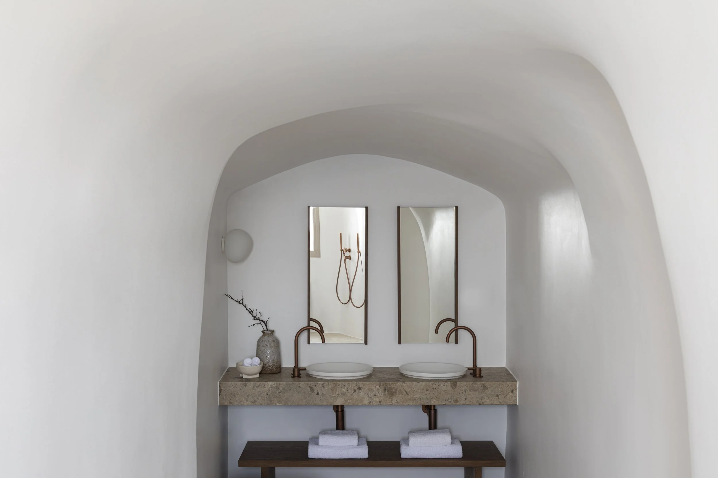 Minimalist bathroom with dual sinks, two mirrors, and a beige stone countertop, featuring a vase with a branch and a bowl of soap.