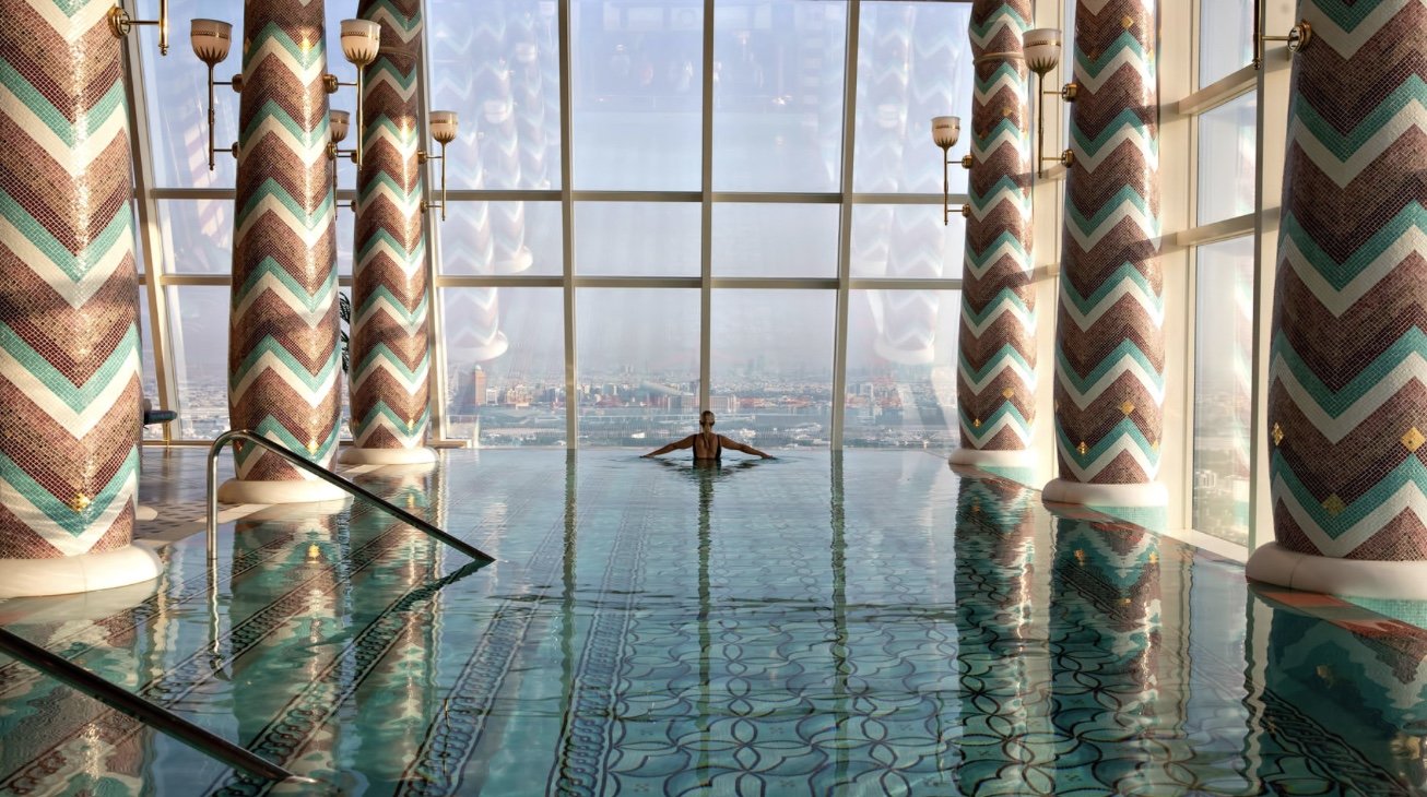 An indoor swimming pool with floor-to-ceiling windows offering a city skyline view. A person is floating in the pool, surrounded by decorated columns and ornate tile flooring.