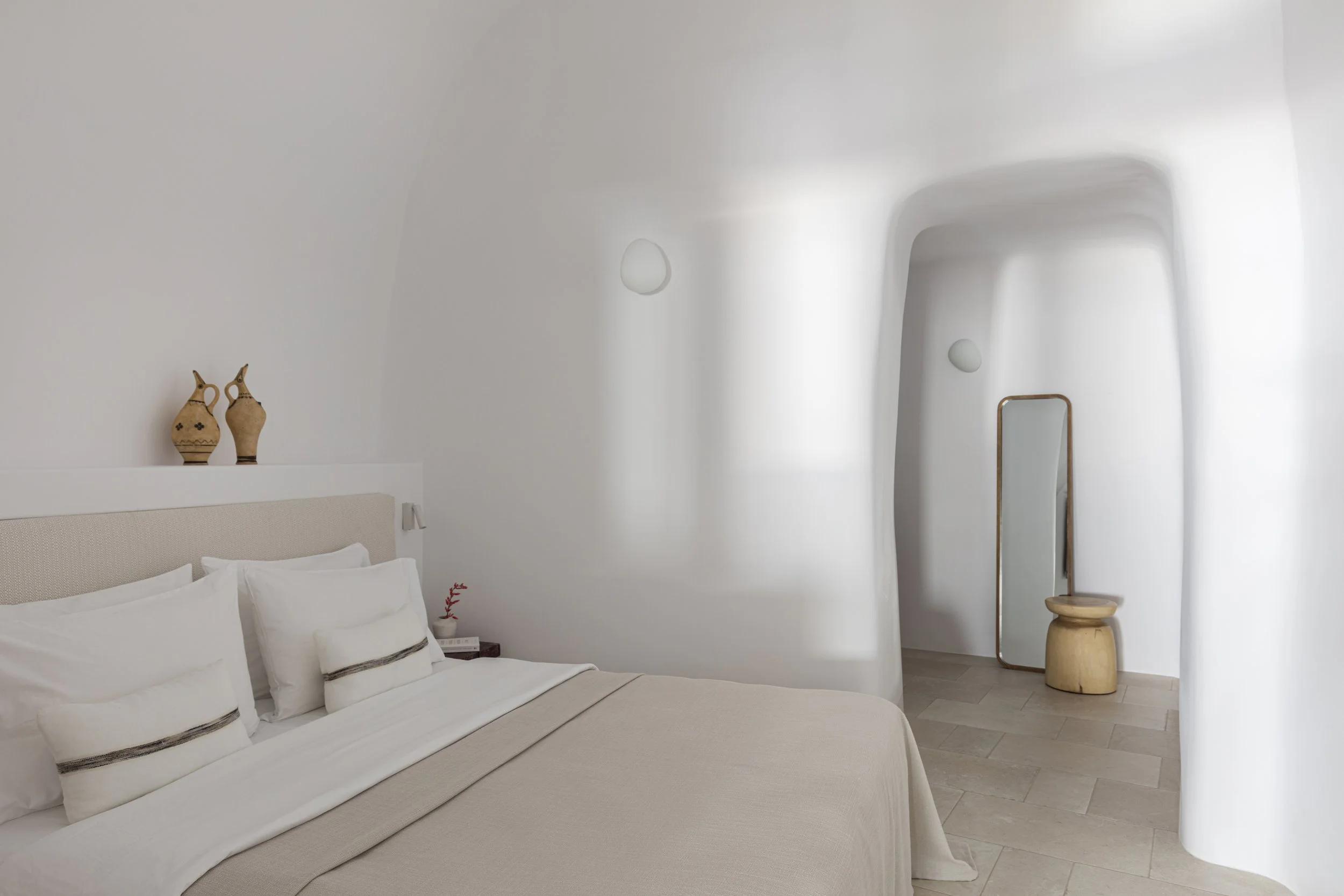 Minimalist bedroom with a white bed, decorative vases on the headboard shelf, a tall mirror, a small wooden stool, and soft lighting from wall-mounted lights with a curved wall in the background.