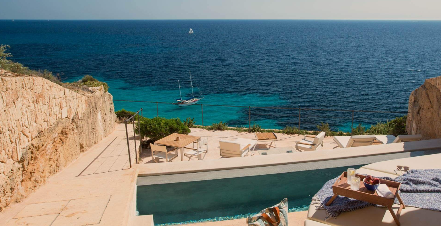 View of an outdoor patio overlooking the ocean with a sailboat in the distance, sun loungers, a table, and a poolside tray with drinks and fruit.