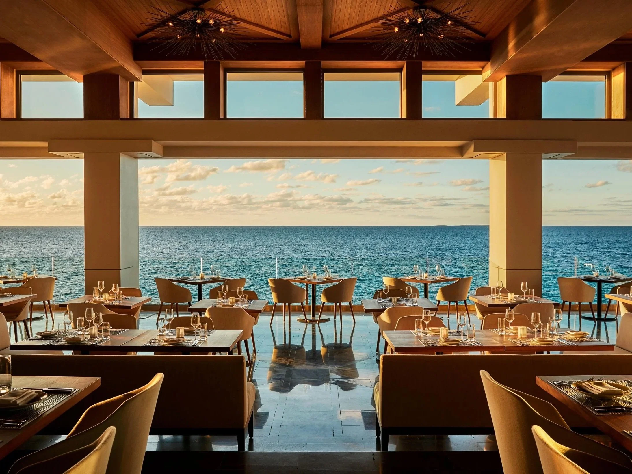 Interior of a restaurant with large open windows overlooking the ocean, tables set with glassware, plates, and napkins, with a sunset sky in the background.