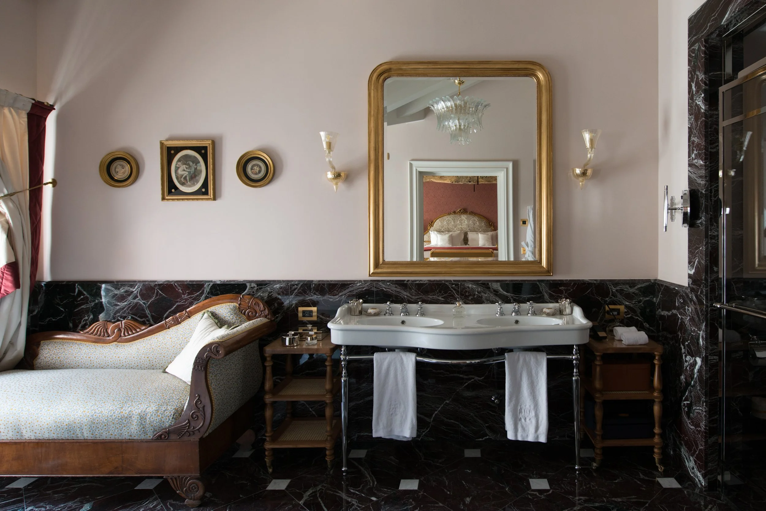 Elegant bathroom with a large mirror over a double sink, vintage wooden chaise lounge on marble floors, and framed artwork on light-colored wall
