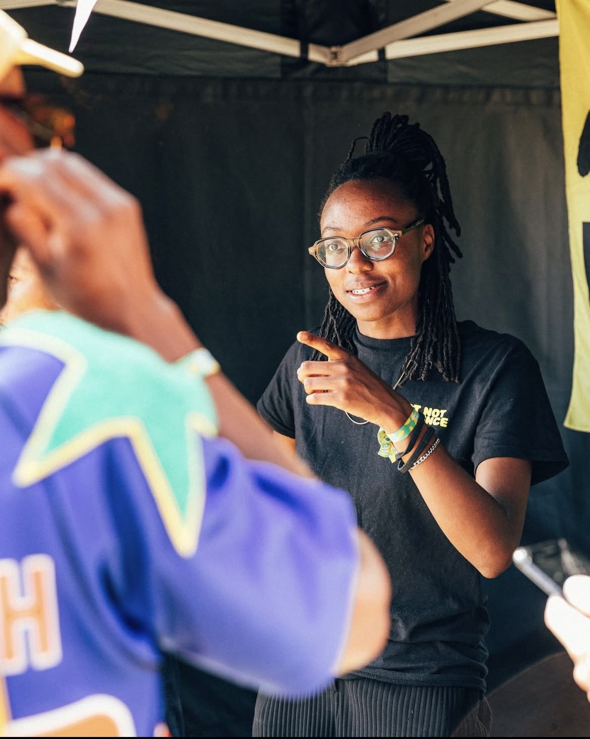 A woman with glasses and dreadlocks pointing and engaging in conversation with another person at an outdoor event, under a tent.