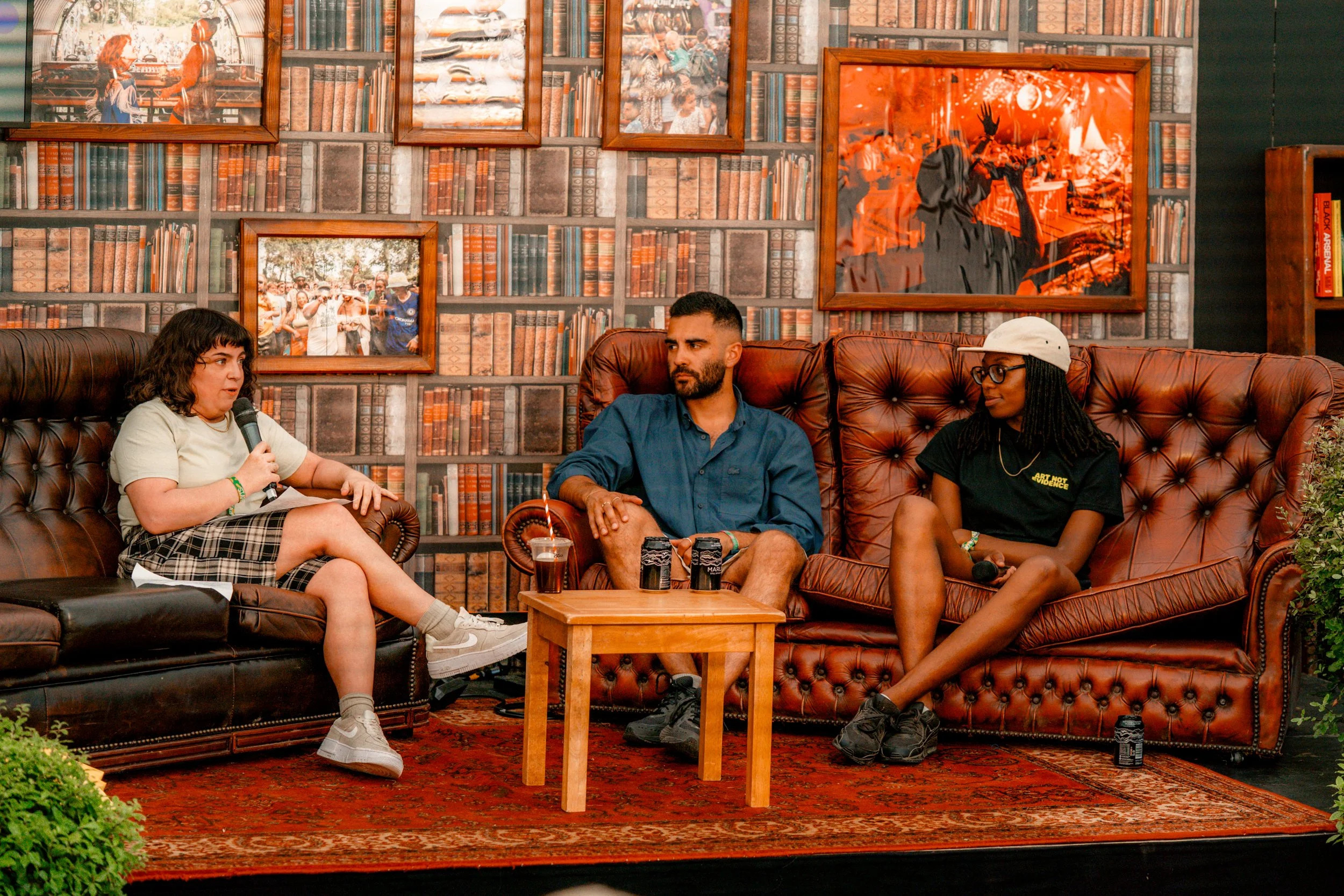 Three people sitting on sofas in a cozy room, engaged in a conversation. The person on the left, a young woman with curly hair, is holding a microphone. The middle person, a man with short dark hair and a beard, is watching her. The person on the right, a woman with glasses and dreadlocks, is holding a microphone. Behind them is a wall decorated with framed pictures and a bookshelf.