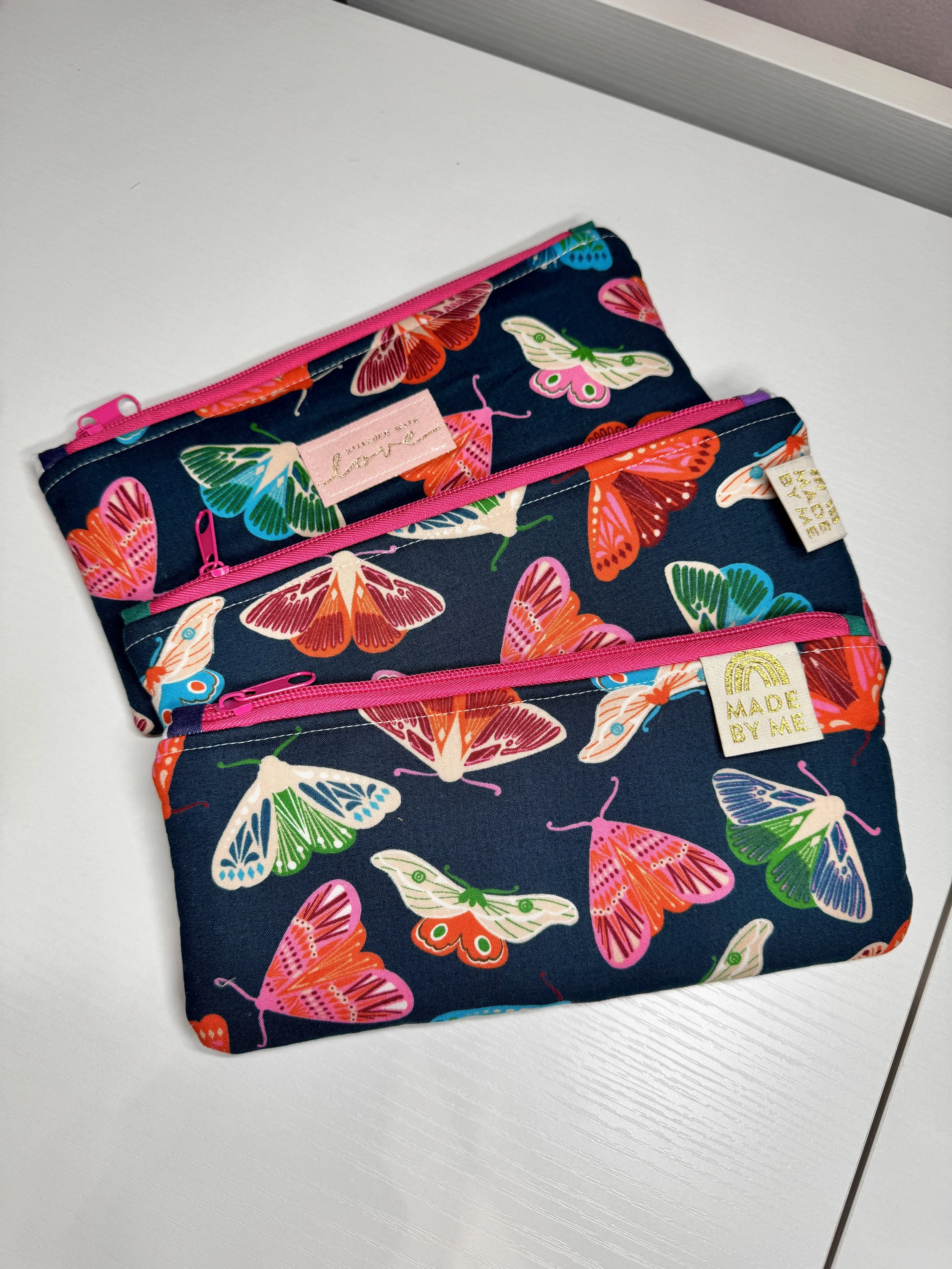 Three fabric zippered pouches with a butterfly pattern on a white surface. Each pouch has a different colored zipper and a small tag with sayings.