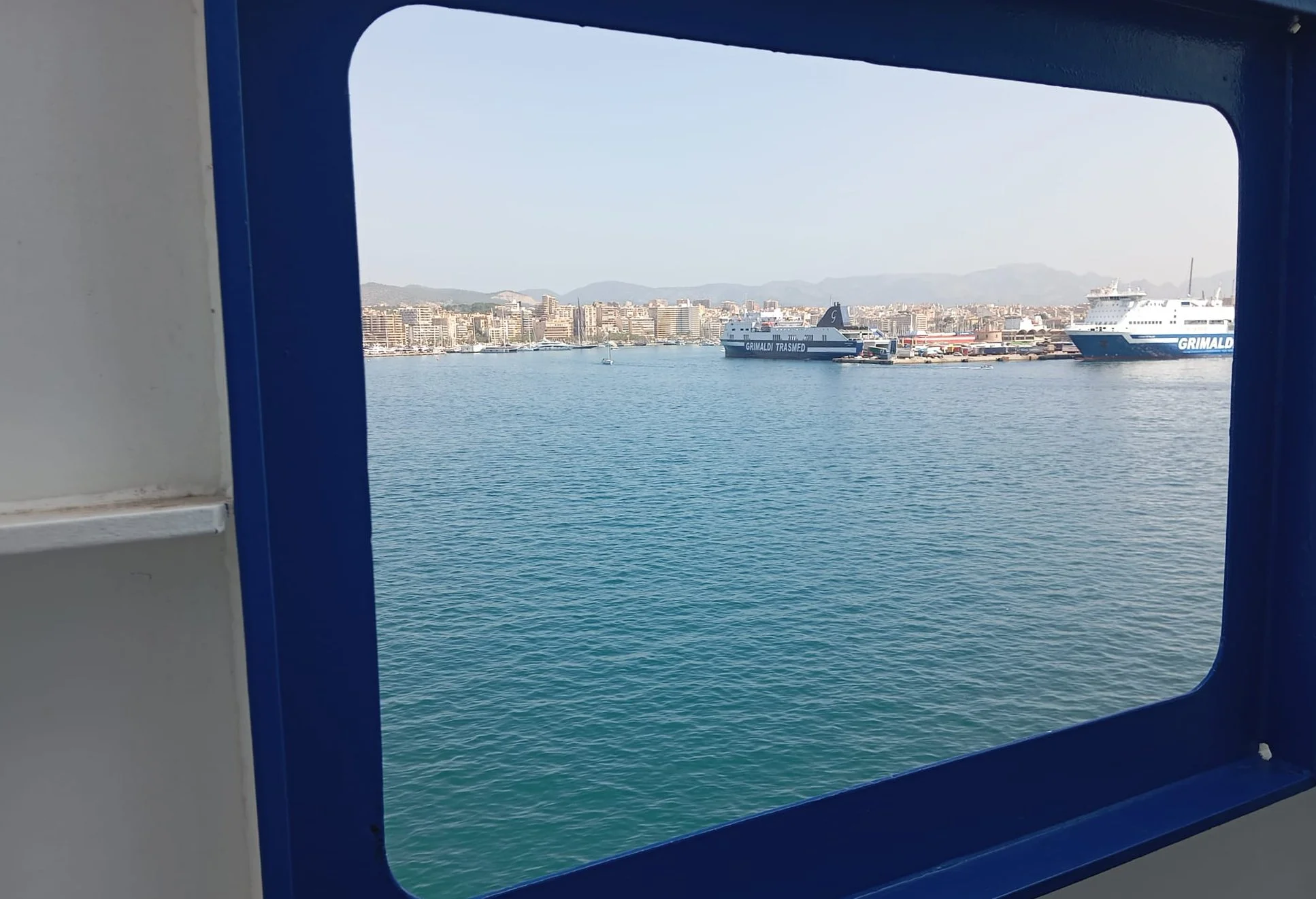 Vista de un puerto con un barco y un ferry en el agua, vista de la ciudad y montañas al fondo, vista desde una ventana.