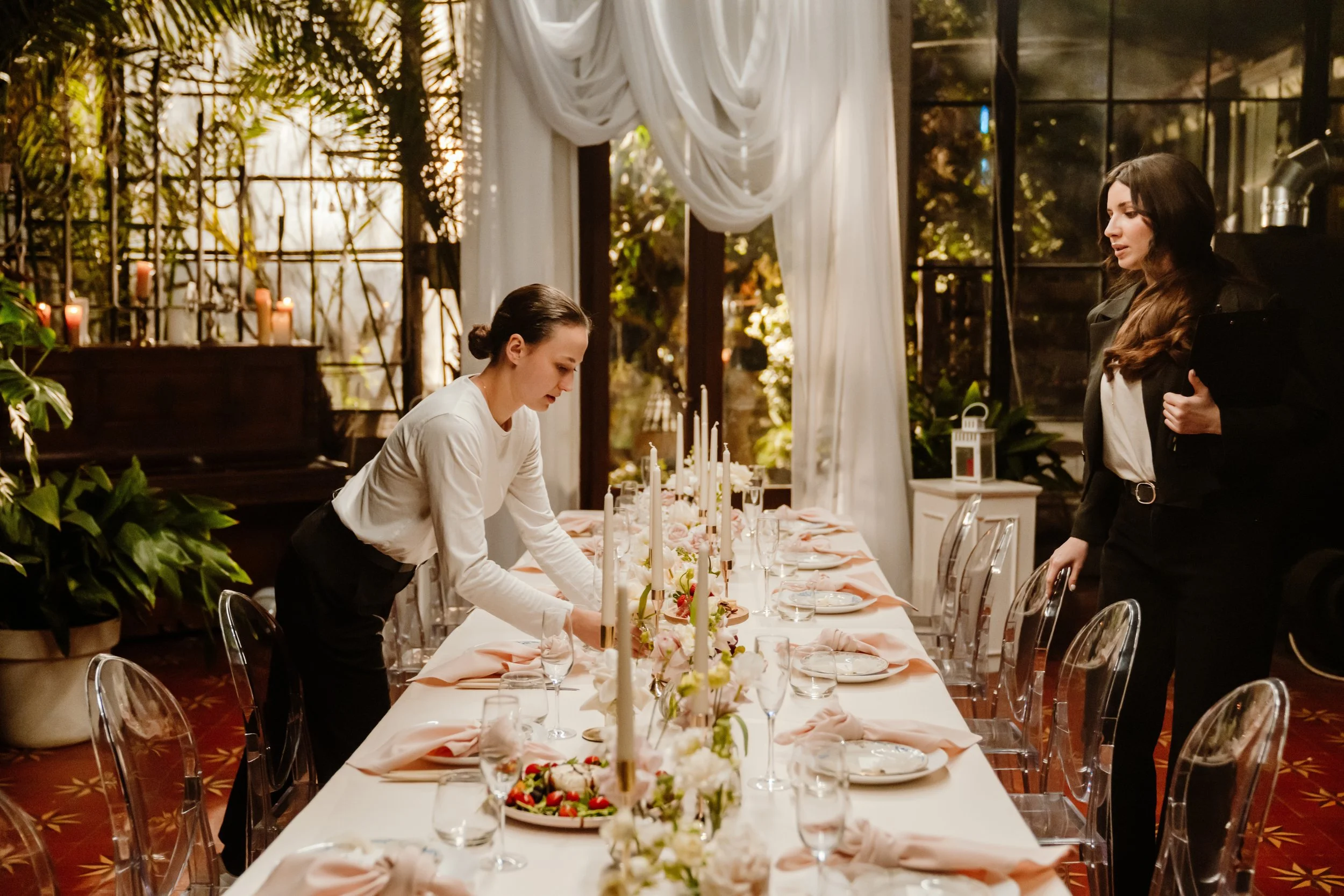 Dos mujeres arreglando una mesa para una celebración en un ambiente elegante y acogedor, decorada con candelabros y flores.