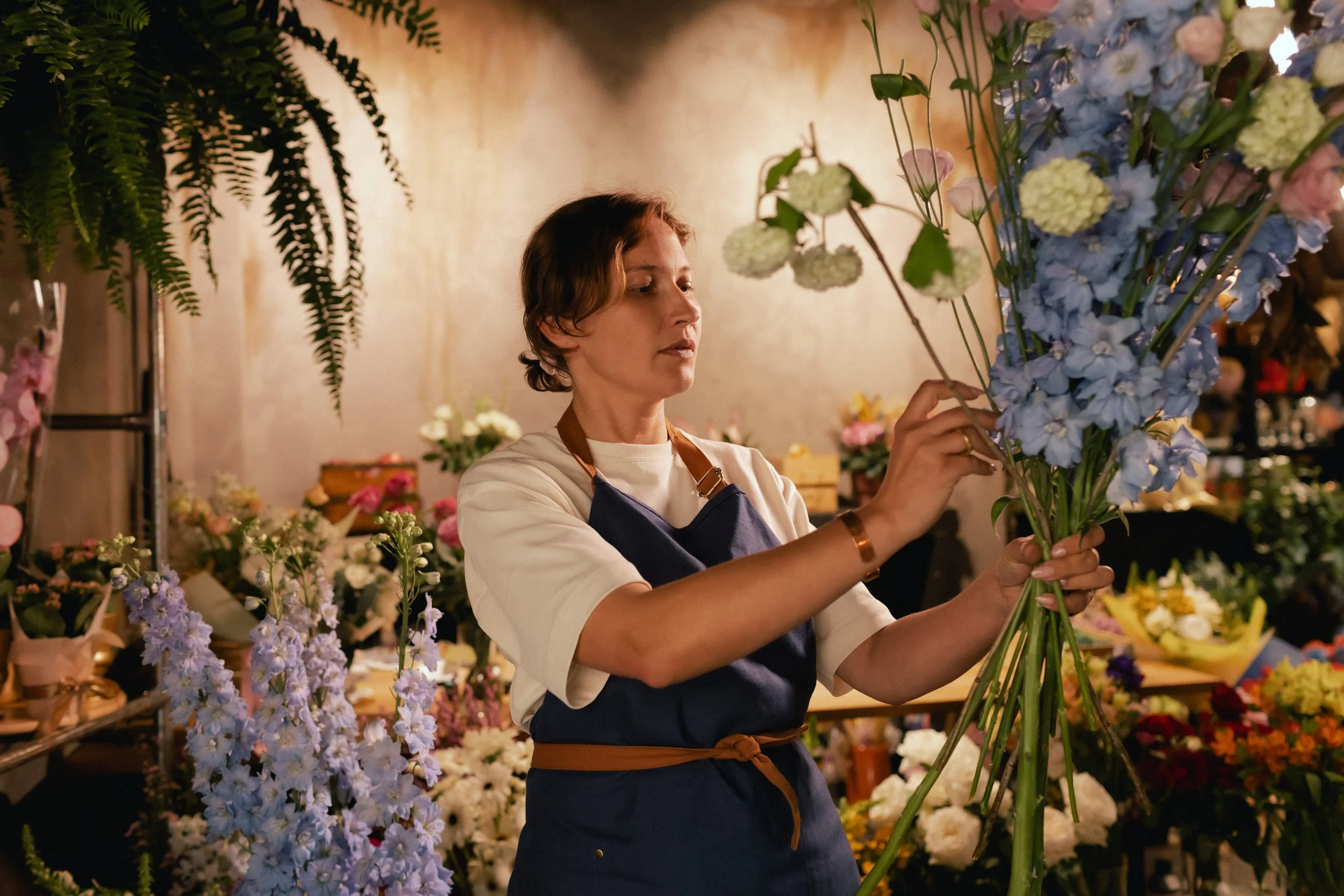 Mujer arreglando flores en un taller de flores, rodeada de arreglos florales de diversos colores y tipos.