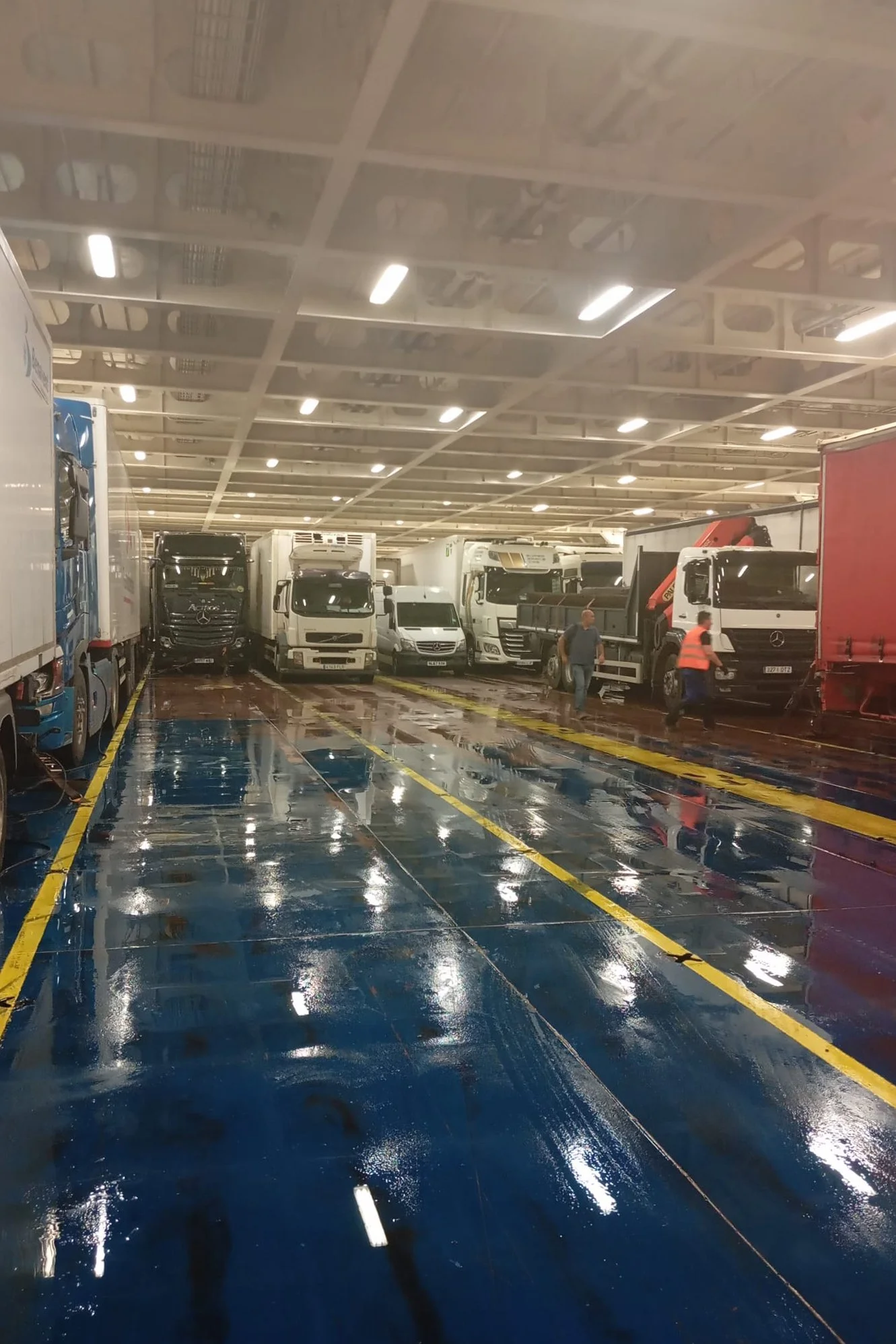 Interior de un ferry con camiones estacionados en la cubierta, piso húmedo y brillante por el agua, con luces blancas en el techo y dos personas caminando.