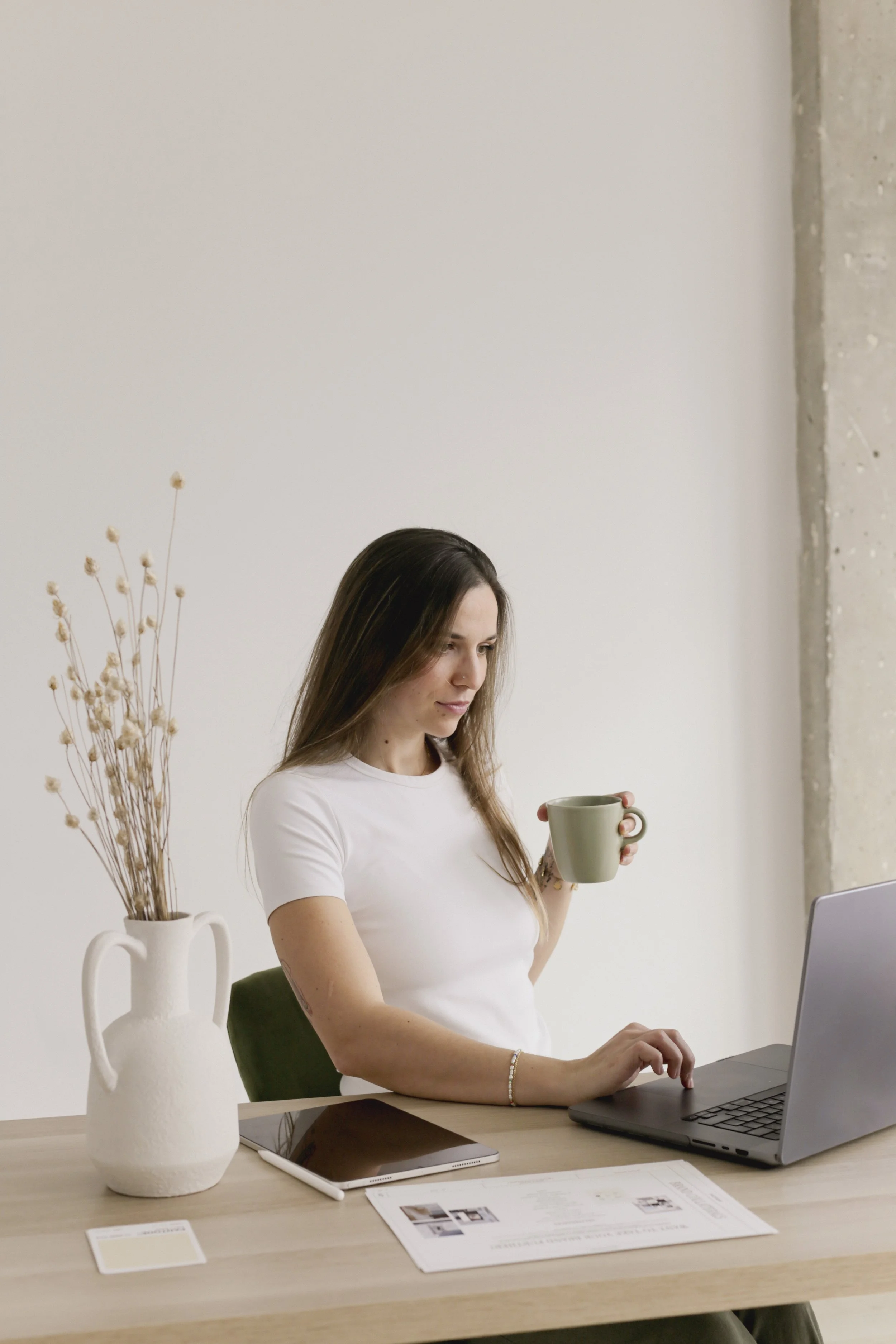 Woman sitting at a desk using a laptop while holding a green mug; a white vase with dry flowers, a tablet, and documents are also on the desk.