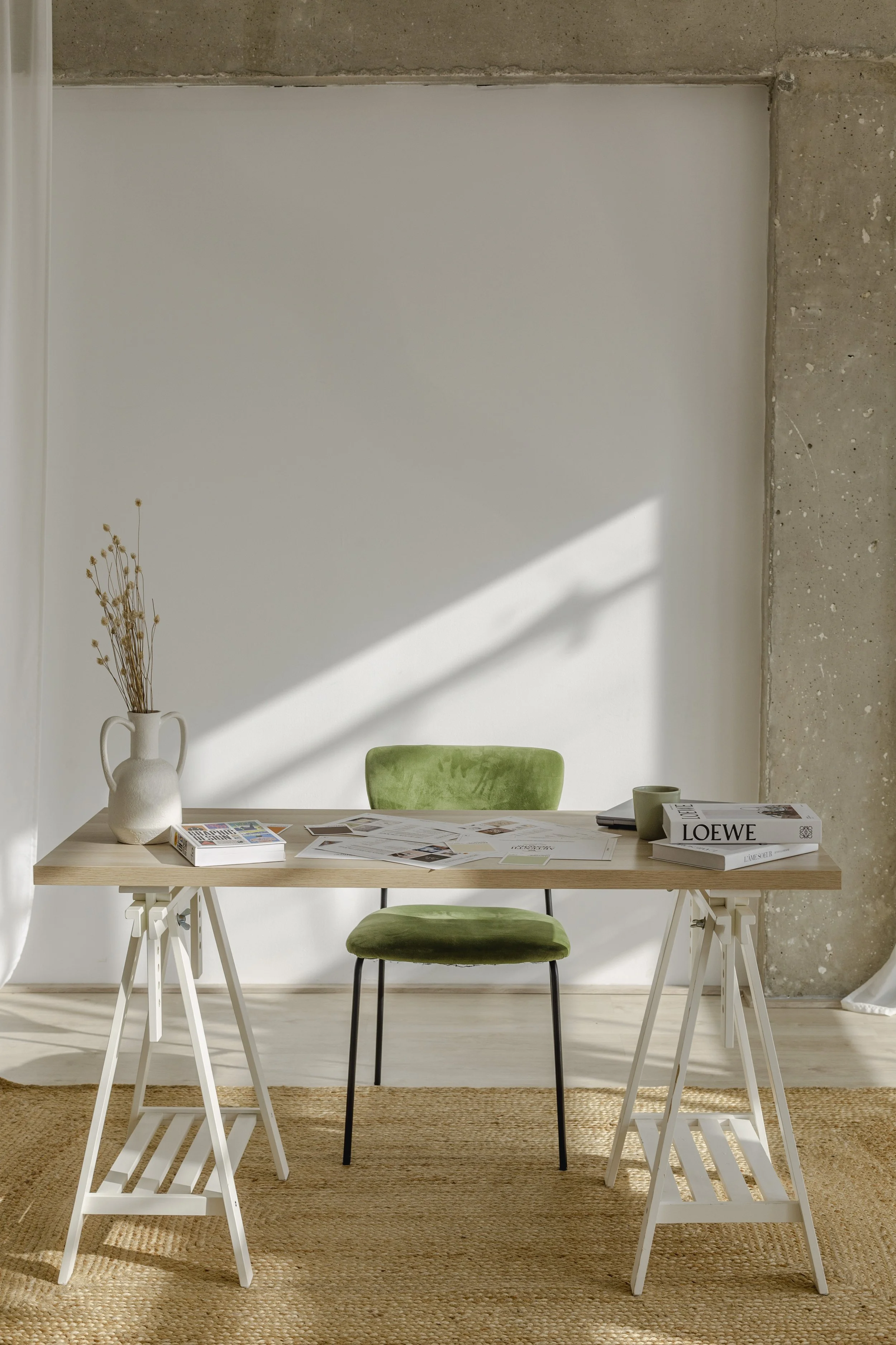 Minimalist office setup with a wooden desk on sawhorses, a green chair, books, papers, and a white vase with dried flowers, against a concrete wall and textured rug.