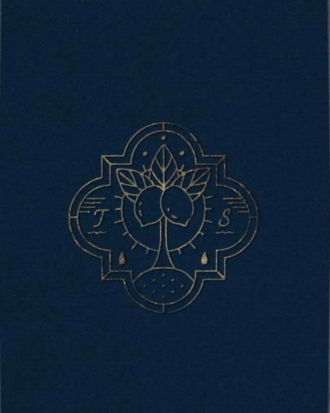 Embossed gold design on dark blue background, featuring a symmetrical illustration of two birds facing each other, surrounded by leaves and decorative elements, with the letters 'T' and 'S' on either side.