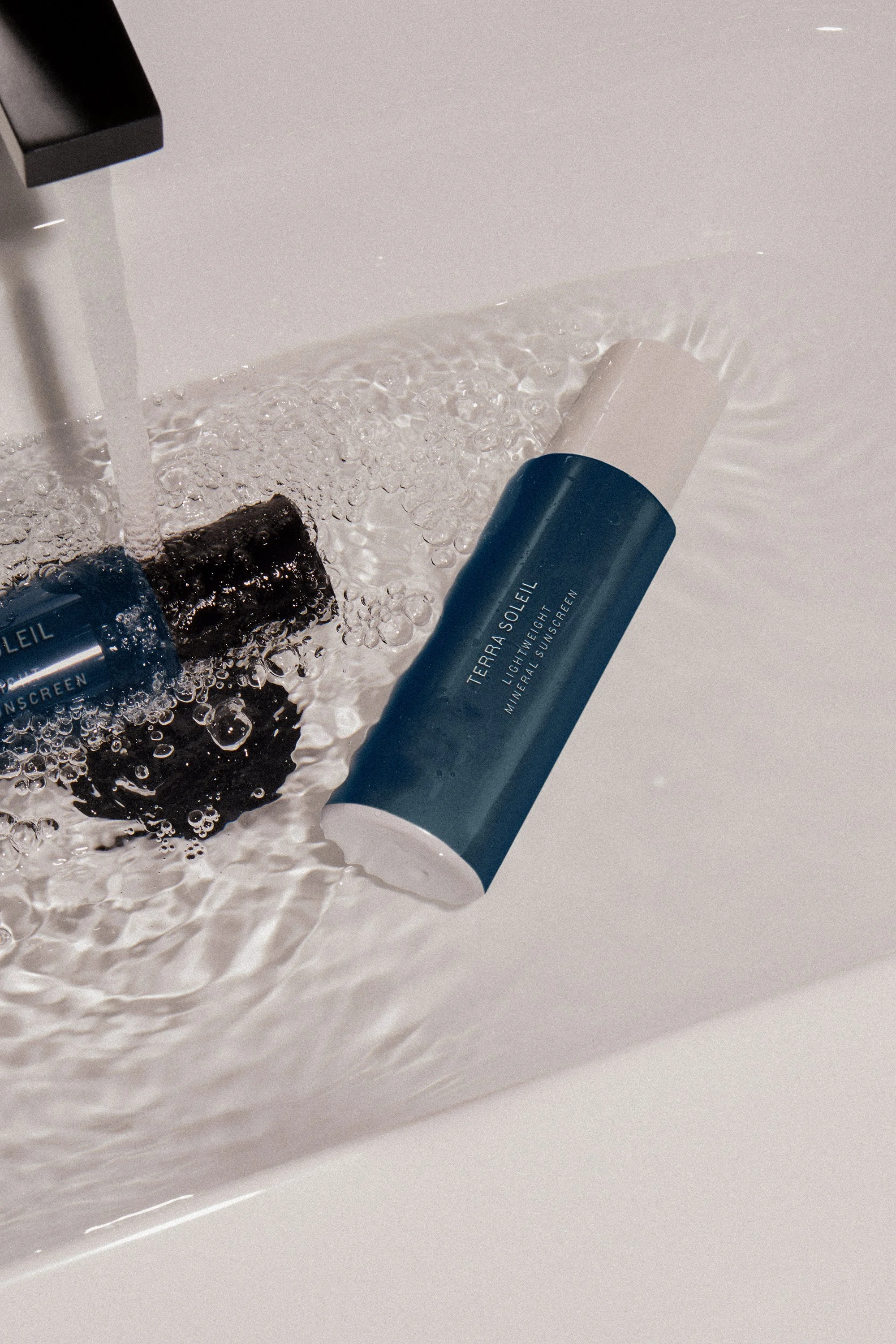 Two blue and white bottles labeled 'Terra Soleil Lightweight Mineral Sunscreen' are under running water in a sink.
