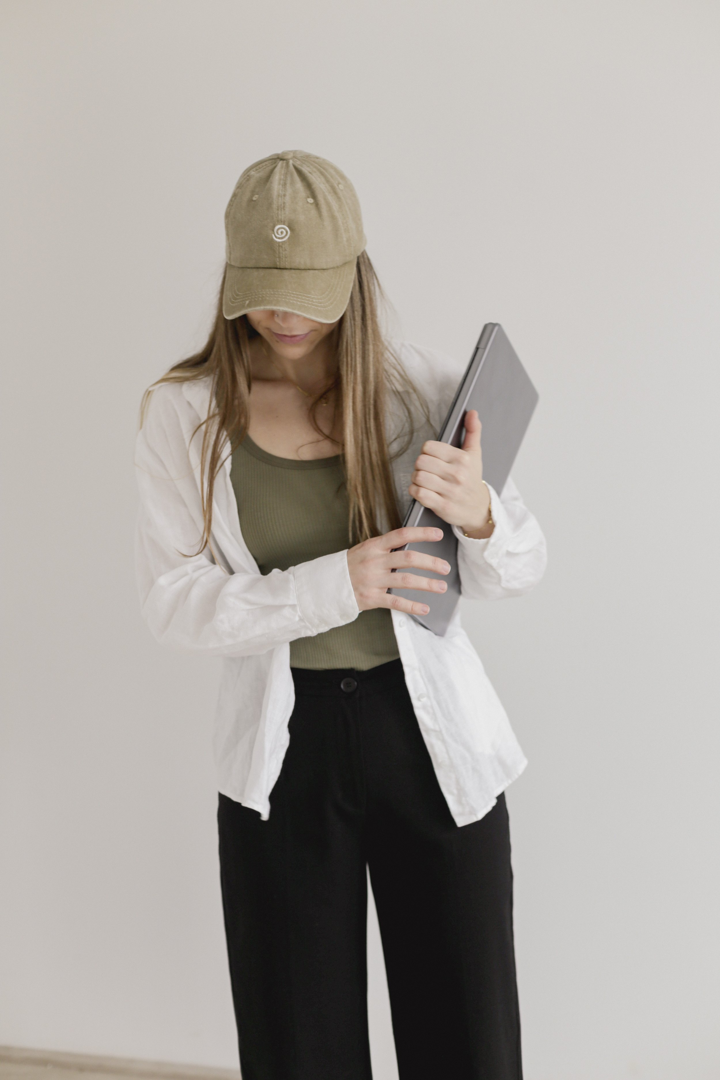 Woman wearing a baseball cap and holding a laptop, dressed in a white shirt and black pants, standing against a neutral background.