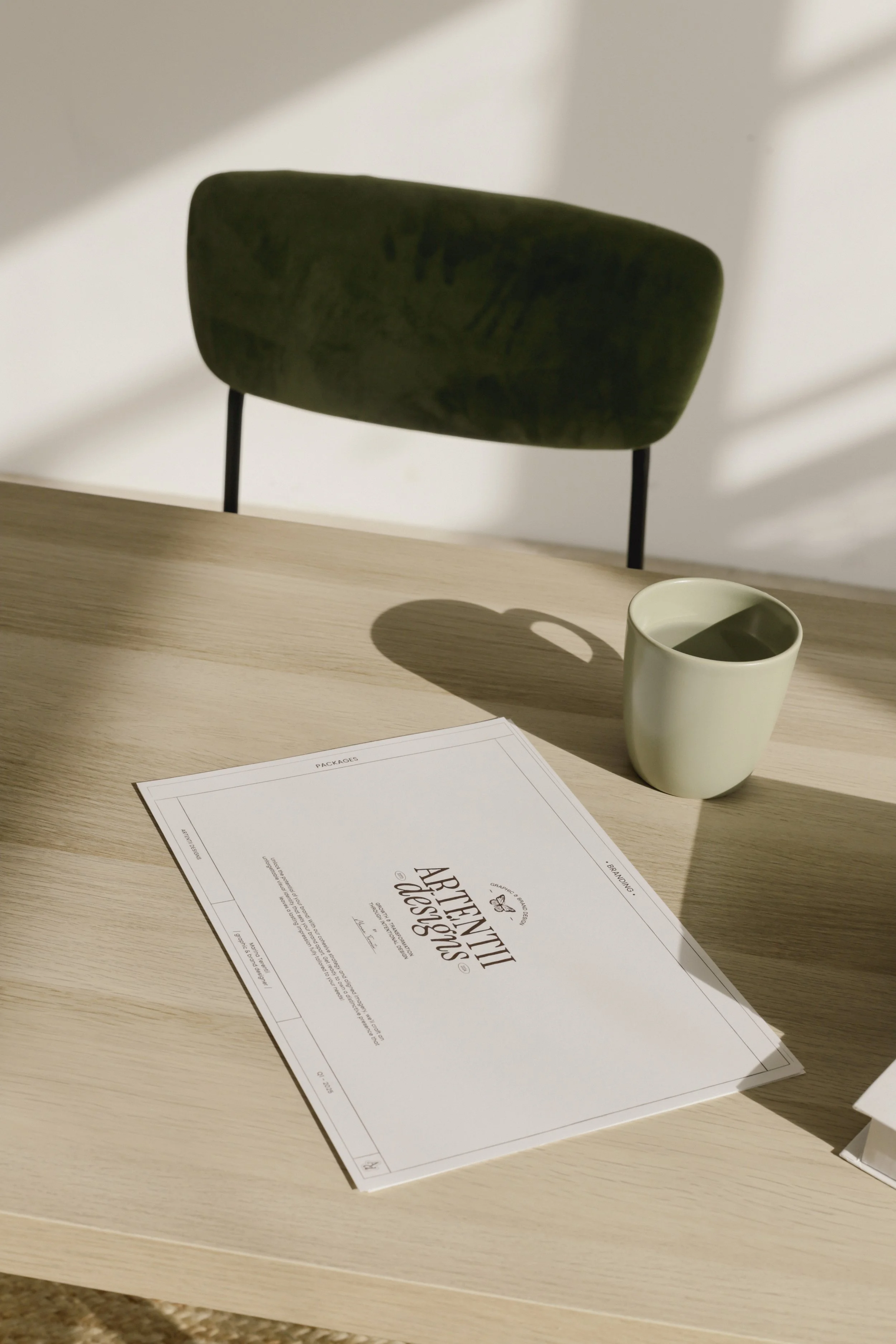 A wooden table with a green chair, a ceramic cup, and a paper with "Artentiti Designs" printed on it in an indoor setting.