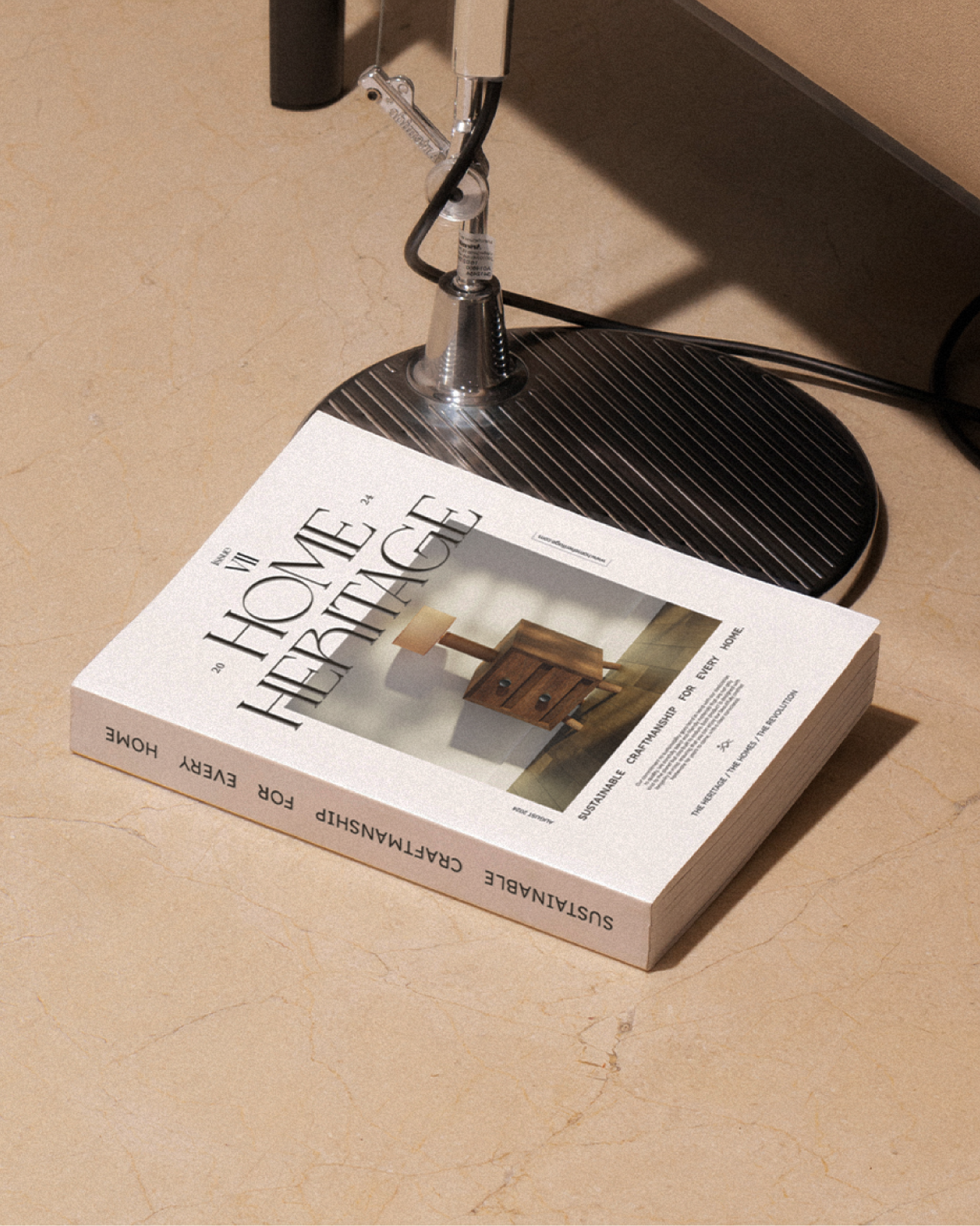 A book titled "Home Heritage" placed on a marble floor next to the base of a chrome lamp. The book focuses on sustainable craftsmanship for home decor, featuring a wooden lamp on its cover.