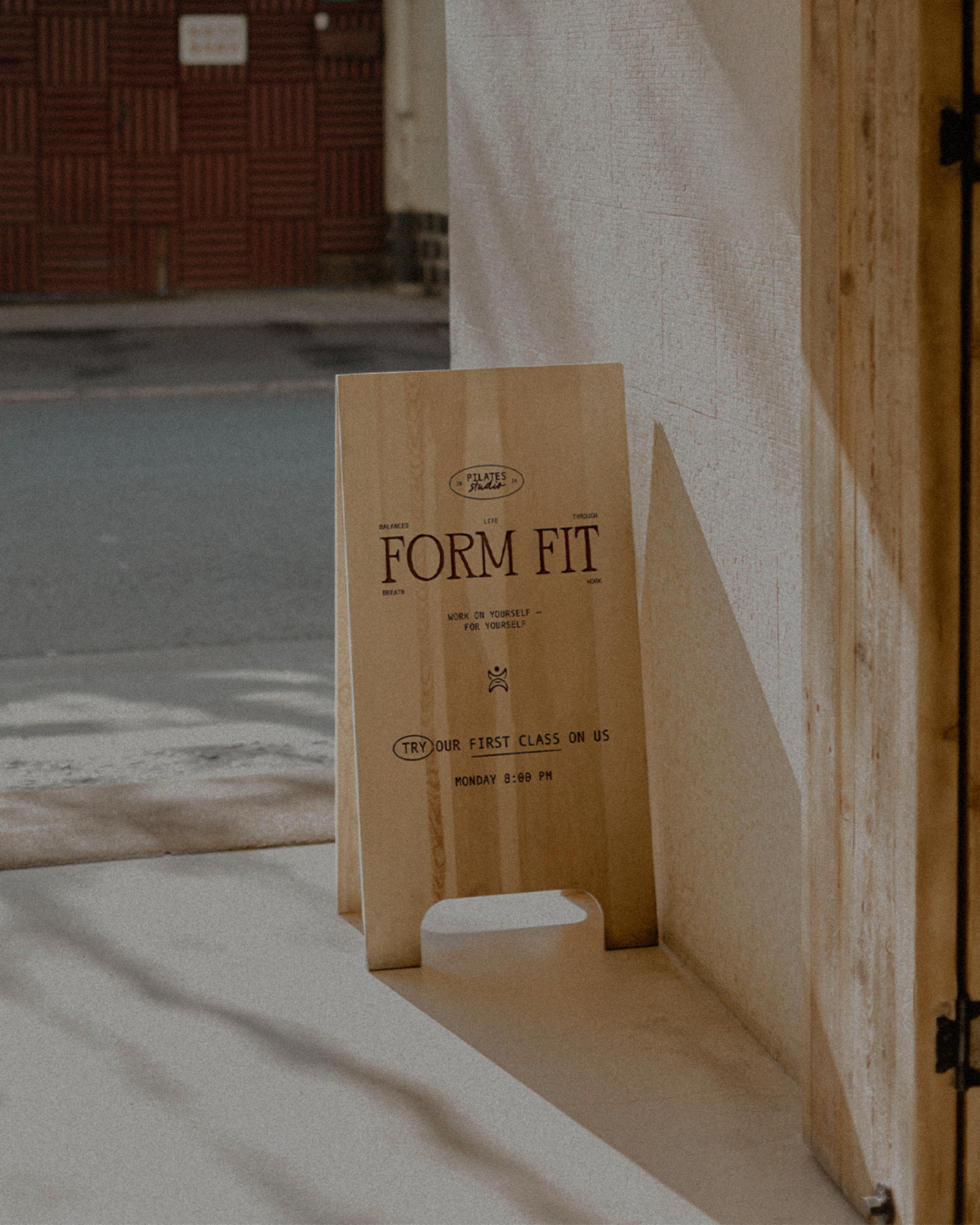 Wooden sandwich board sign with text advertising "Form Fit," offering a free first class on Pilates. Sign is on a sidewalk near a building entrance.