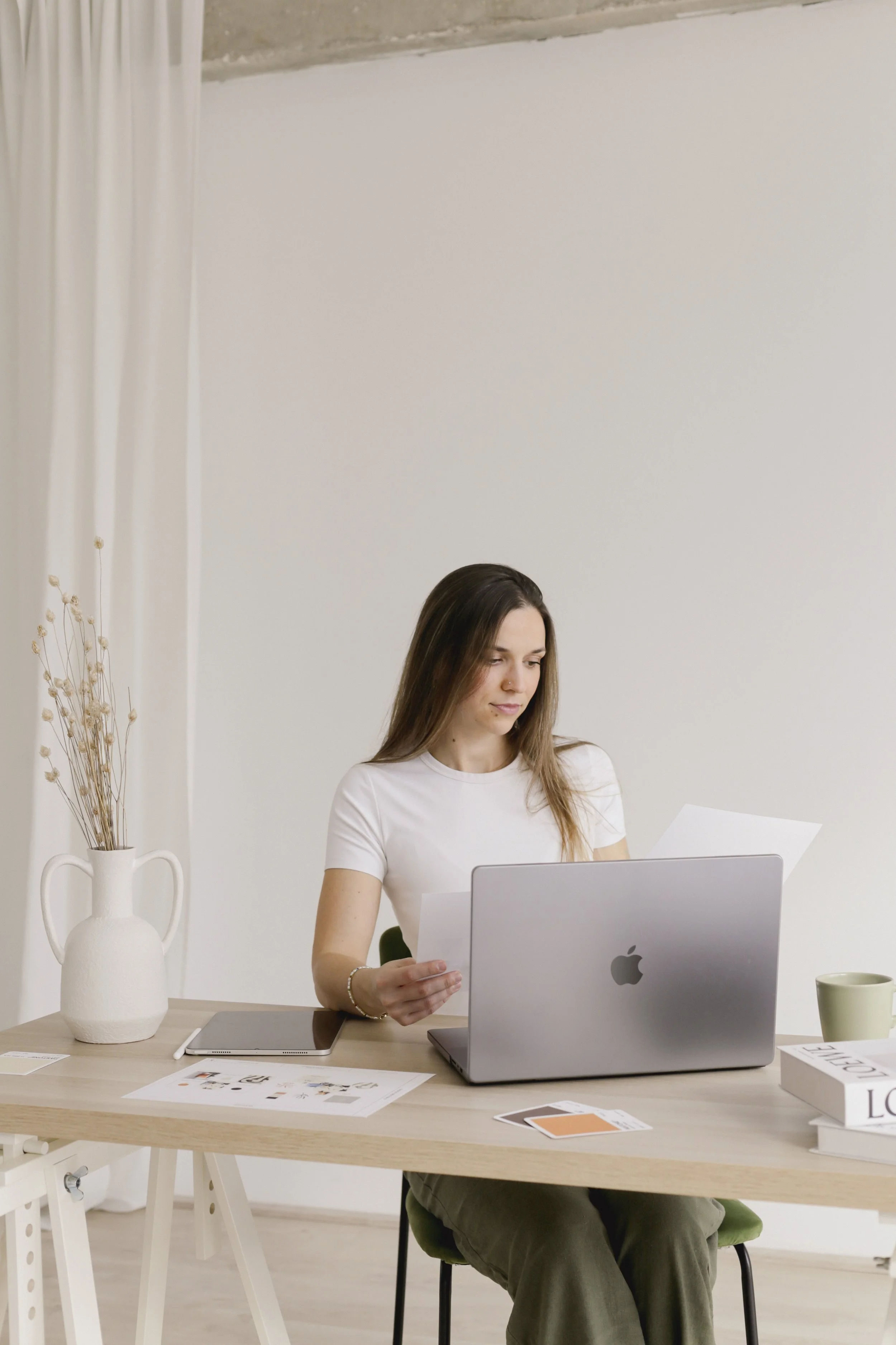 Woman sitting at a desk with a laptop, holding documents, and a vase with dried flowers.