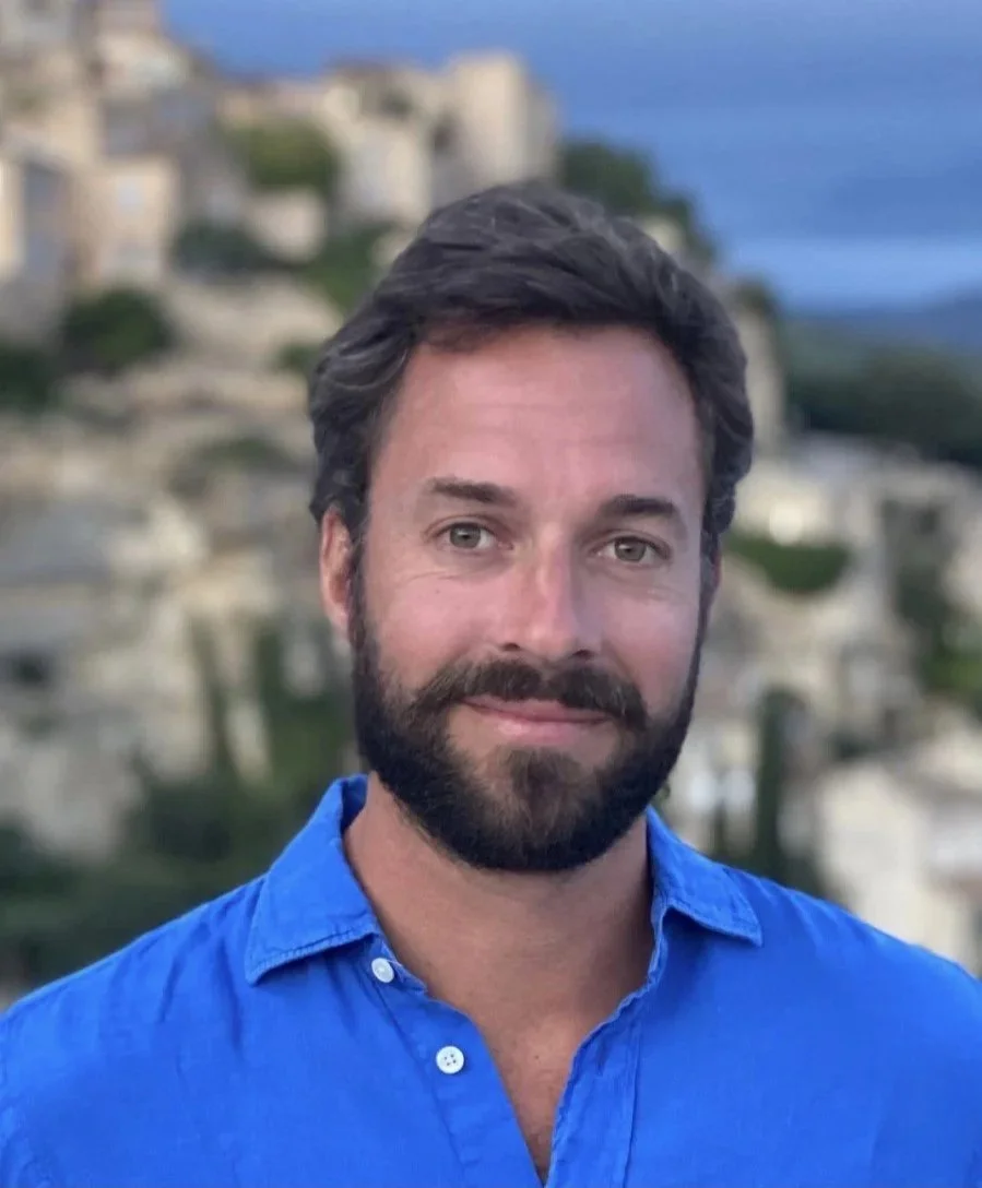 A man with dark hair and a beard, wearing a blue shirt, standing outdoors with a hilly town and sky in the background.